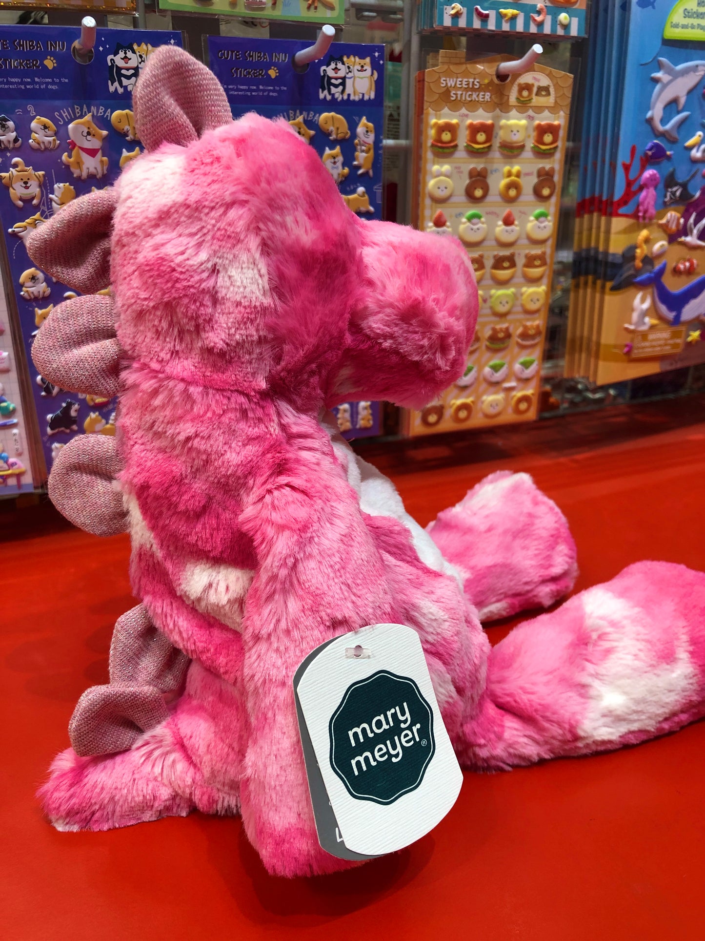 Pink Mary Meyer plush dinosaur toy on display with colorful sticker sets in background, soft and cuddly Marshmallow Zoo character