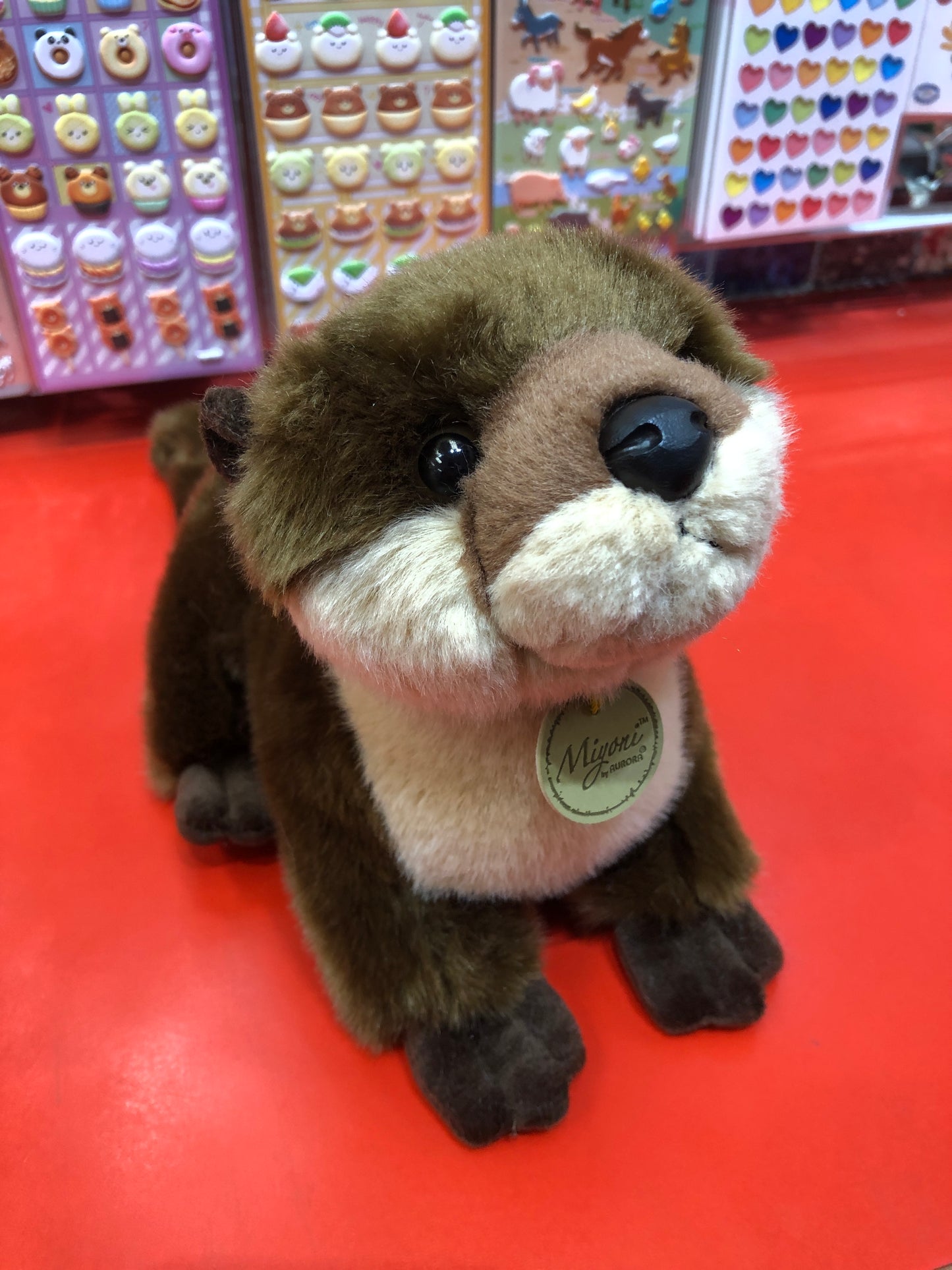 Adorable Aurora Miyoni river otter plush toy on red surface, with colorful sticker display in background