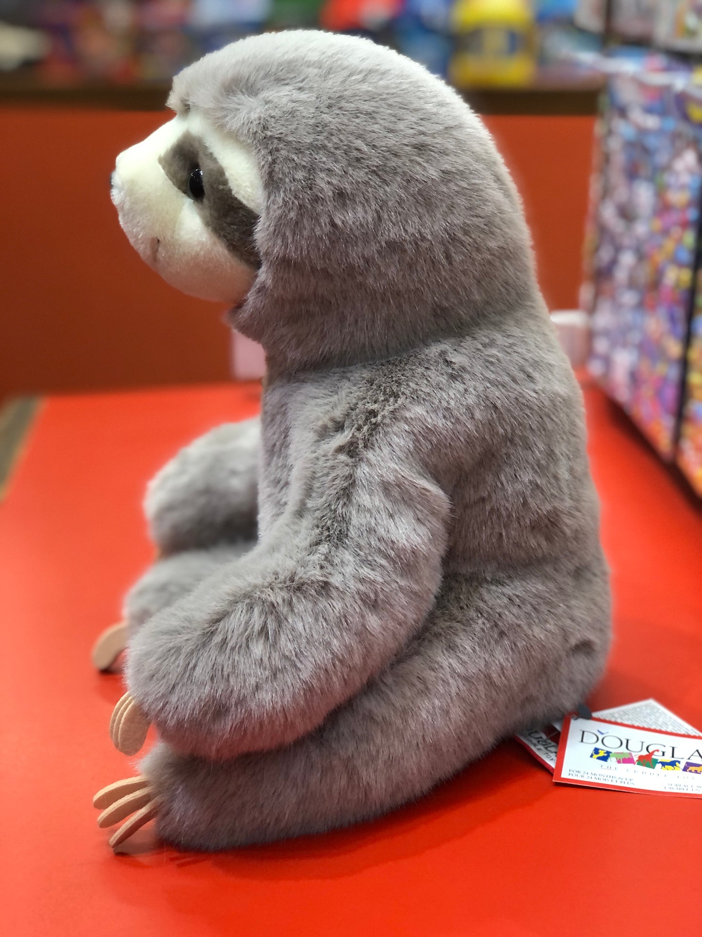 Douglas Super Lizzie Soft Sloth plush toy, gray with white face, sitting on red surface, lifelike features and felt claws