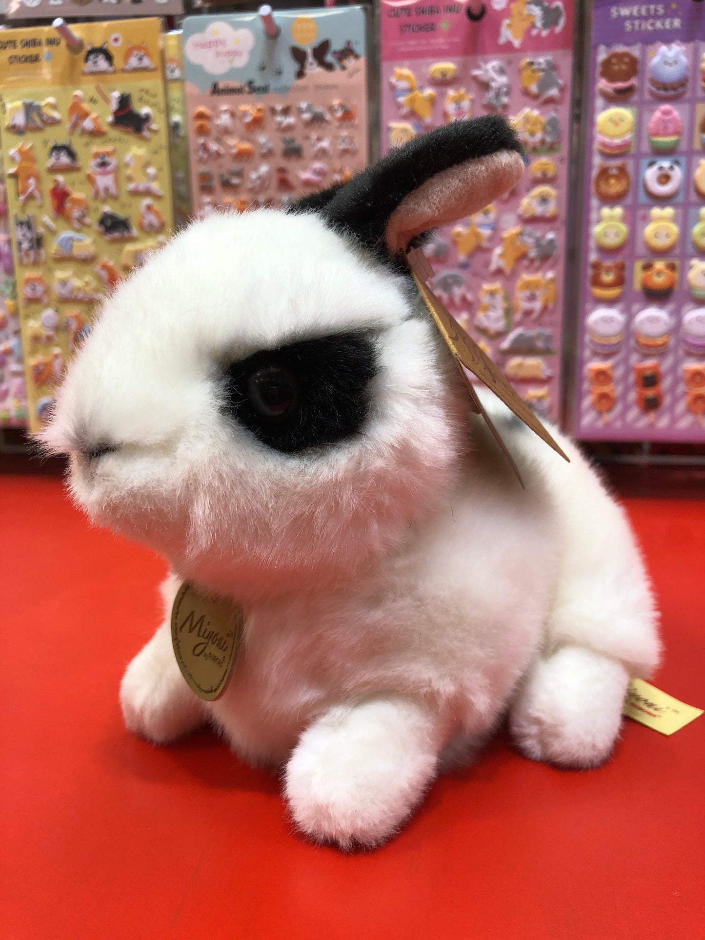 Adorable black and white Rex Rabbit plush toy with pink ears, displayed on red surface with colorful sticker sheets background