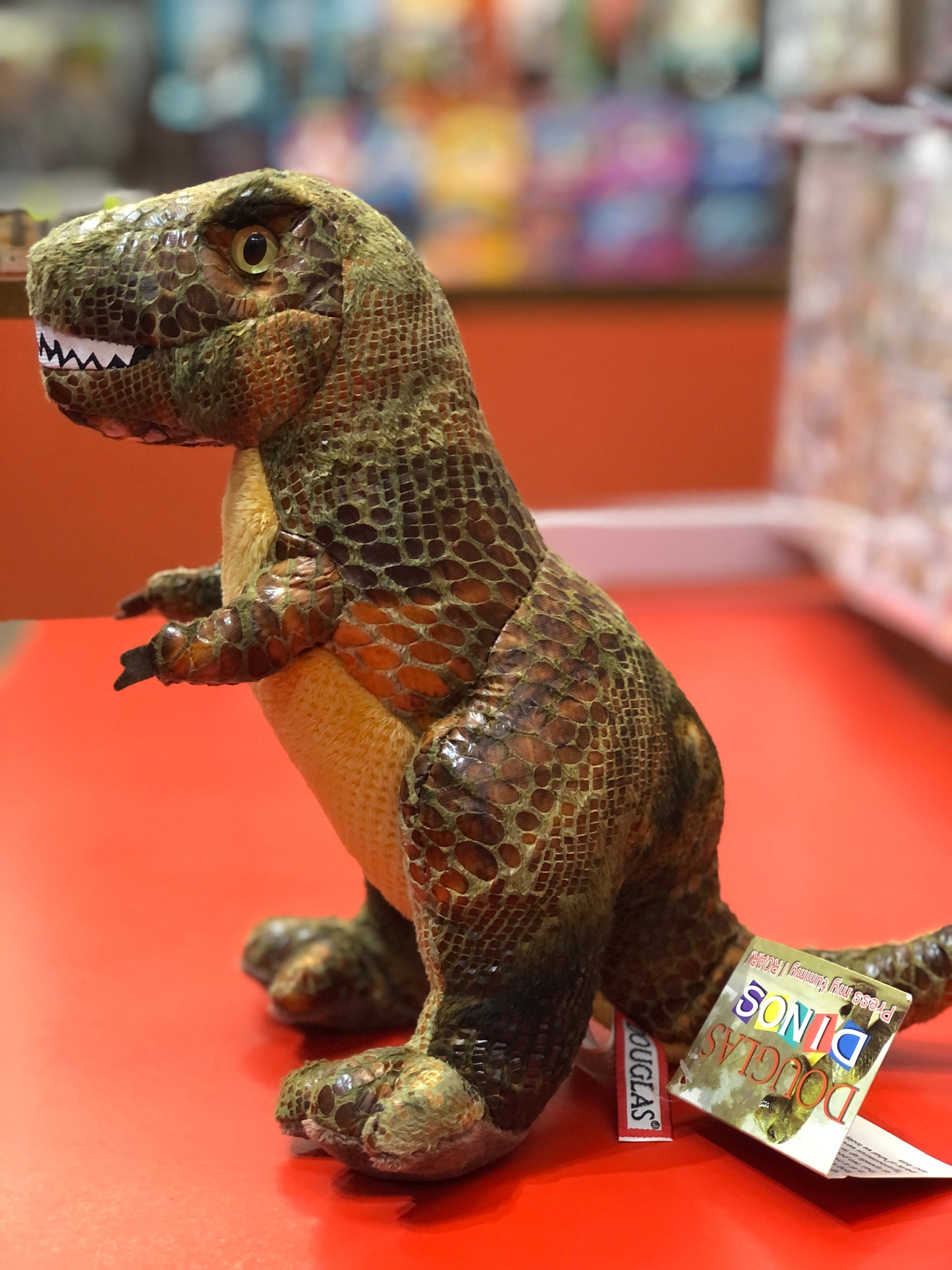Plush T-Rex dinosaur toy with realistic scaly texture and teeth, sitting on red surface with Douglas tag visible