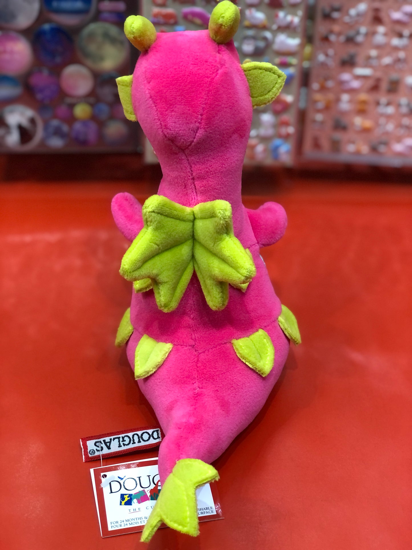 Bright pink Douglas dragon fruit plush toy with green accents, shaped like a cute dragon, sitting on red surface