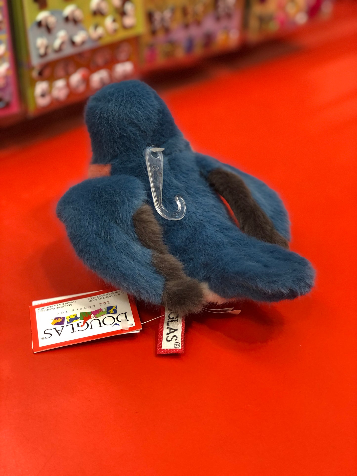 Cuddly blue bird plush toy Bertie on red background, with tag and plastic hook visible, part of Douglas brand