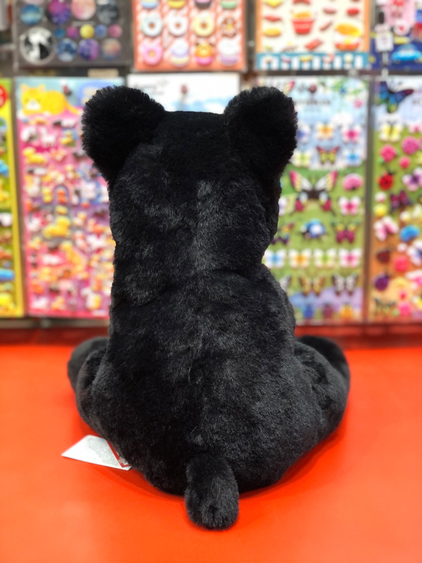 Cuddly black bear plush toy sitting on red surface, colorful toy displays in background, soft and huggable forest animal stuffed animal