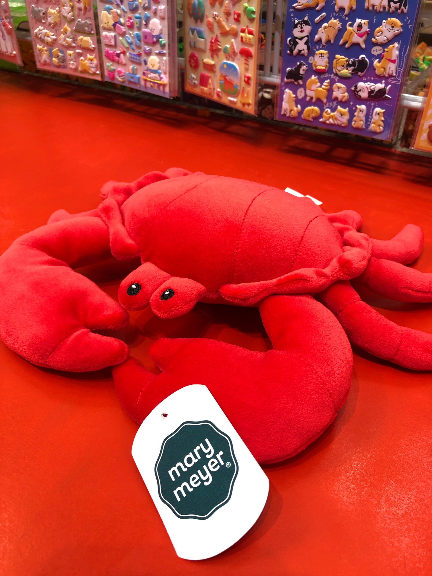 Soft, squishable Mary Meyer Smootheez crab plush toy in bright red, displayed on red surface with colorful stickers in background