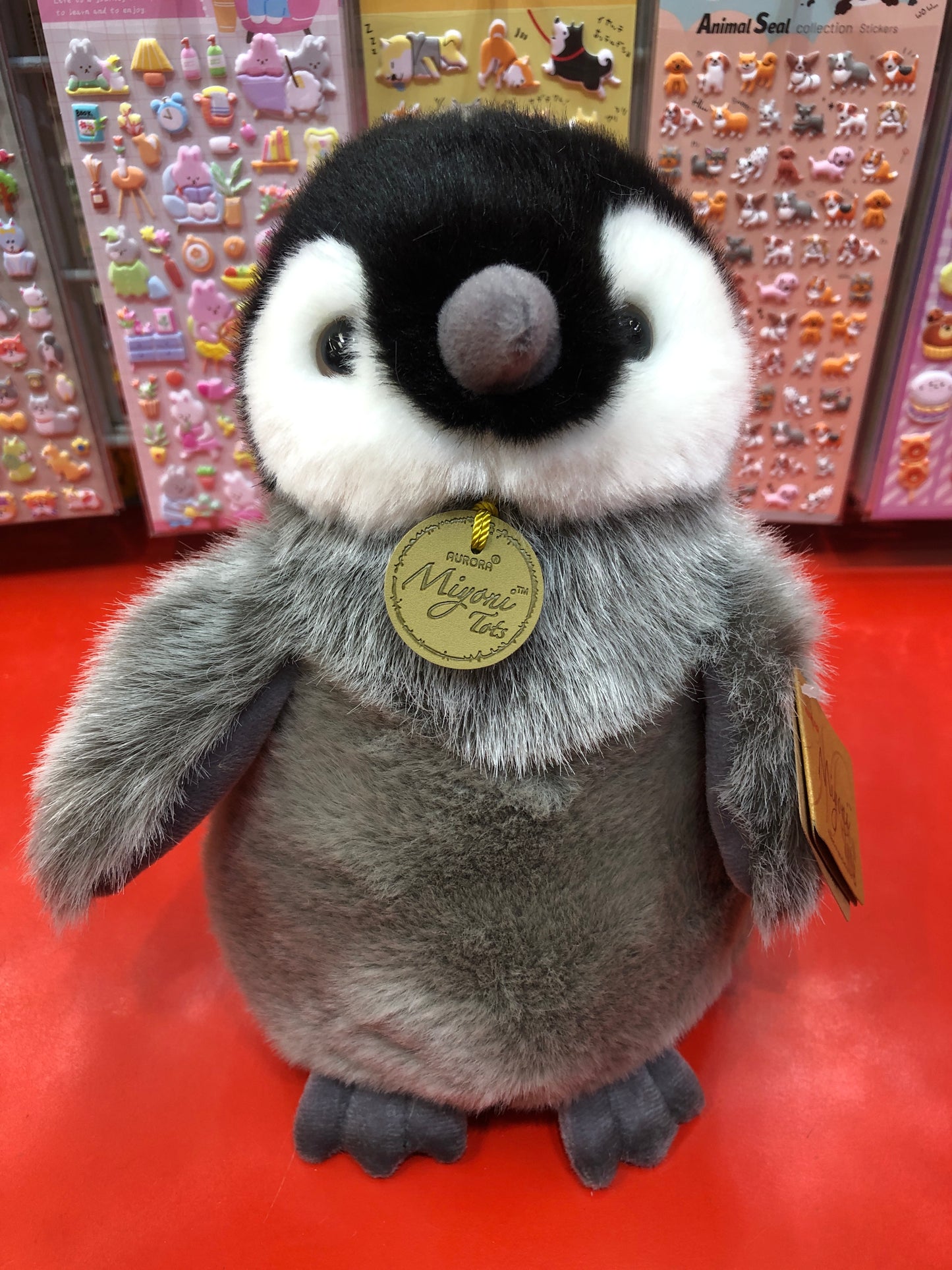 Adorable Aurora Miyoni Tots Emperor Penguin Chick plush toy with fluffy gray feathers and distinctive facial markings on red display