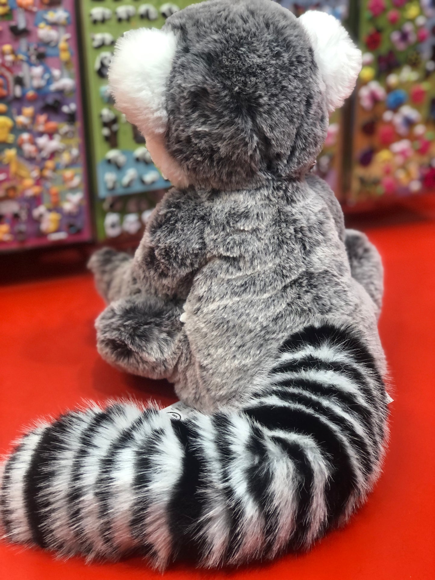 Cuddly gray lemur plush toy with realistic features and striped tail, displayed on red surface with colorful background