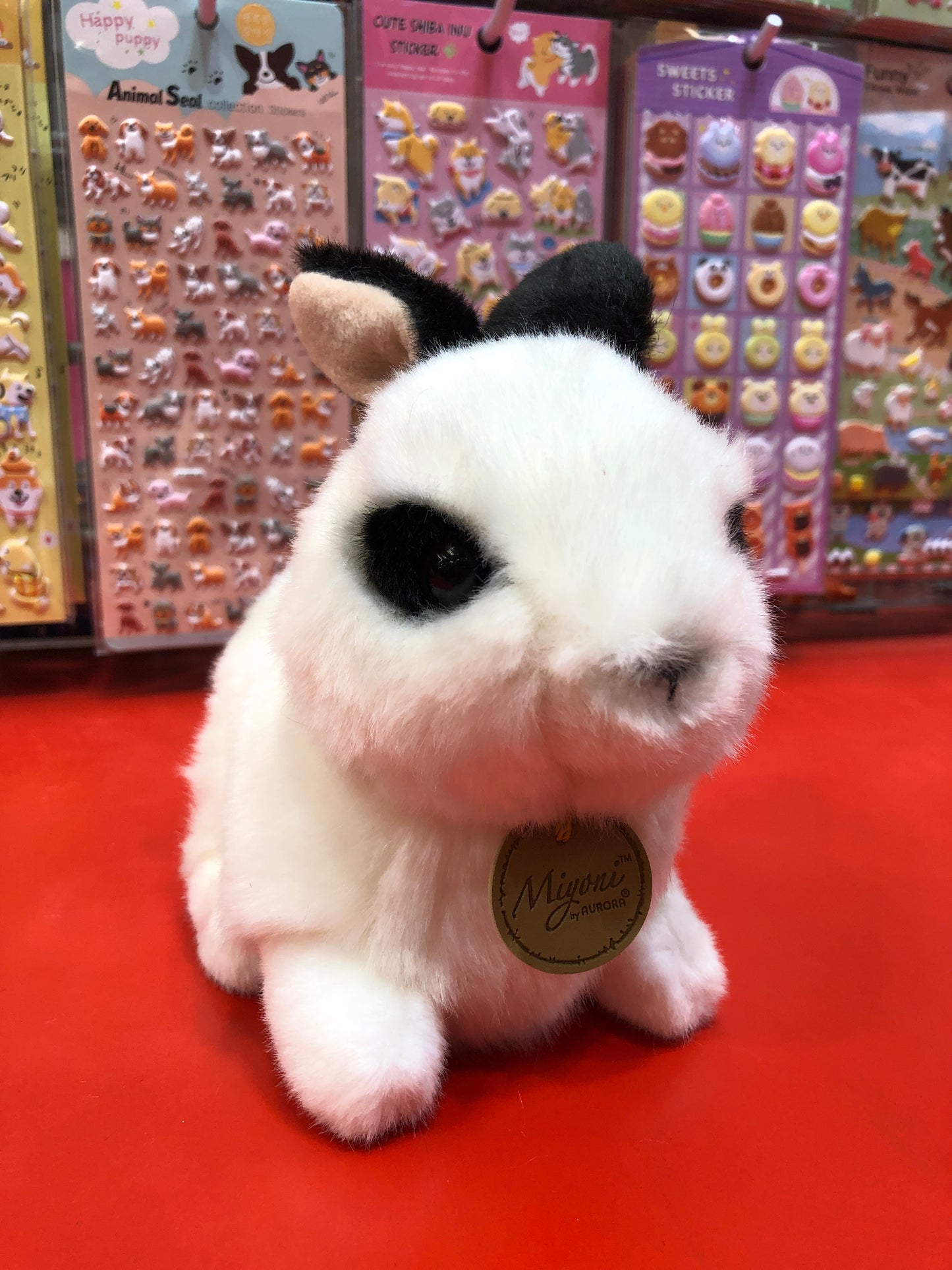 Cute Aurora Miyoni plush Rex Rabbit with black and white fur, sitting on red surface near colorful sticker displays