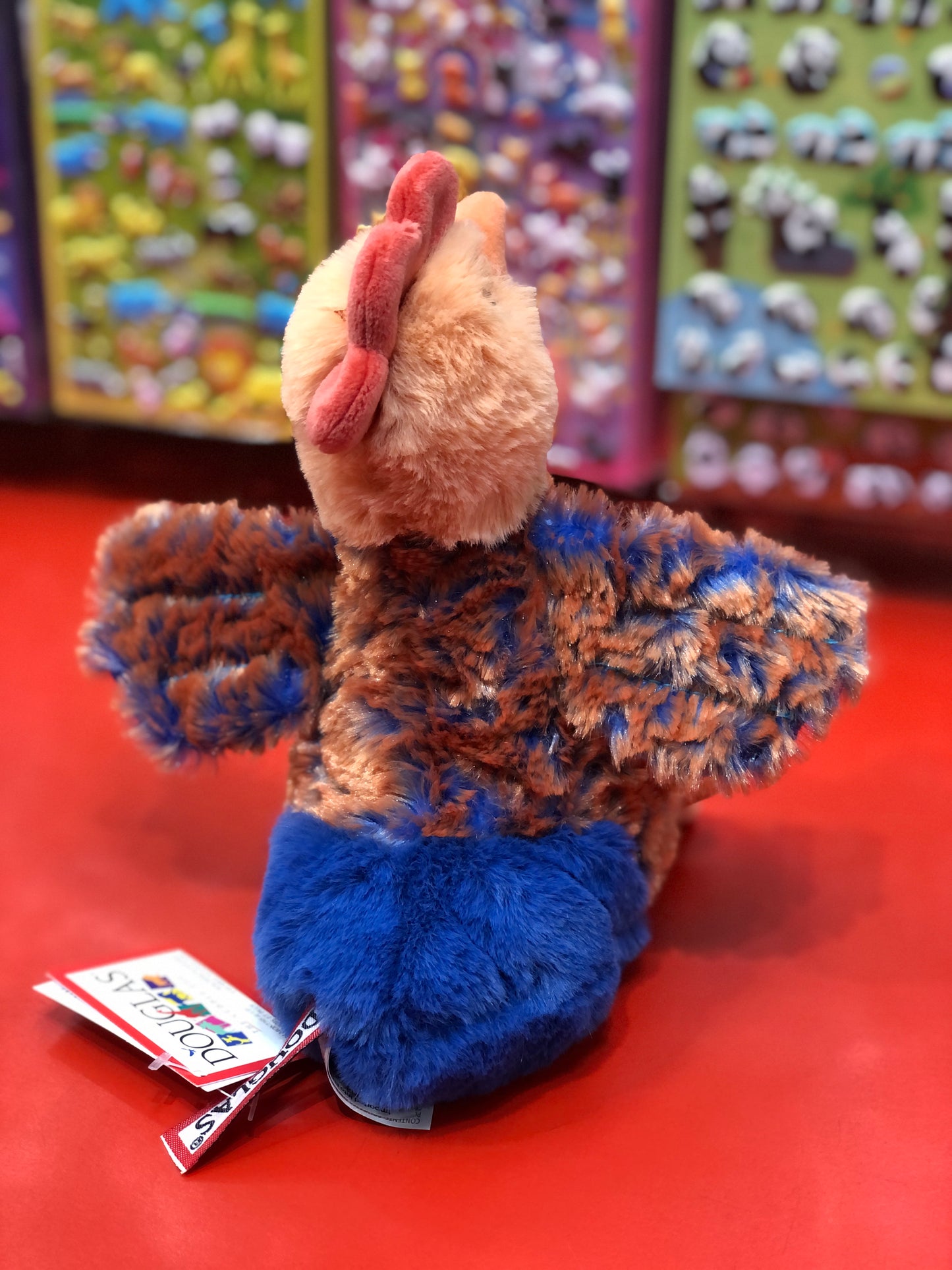 Plush Douglas Chantillie Blue Lace Hen toy with soft blue and brown feathers, displayed on red surface with colorful background
