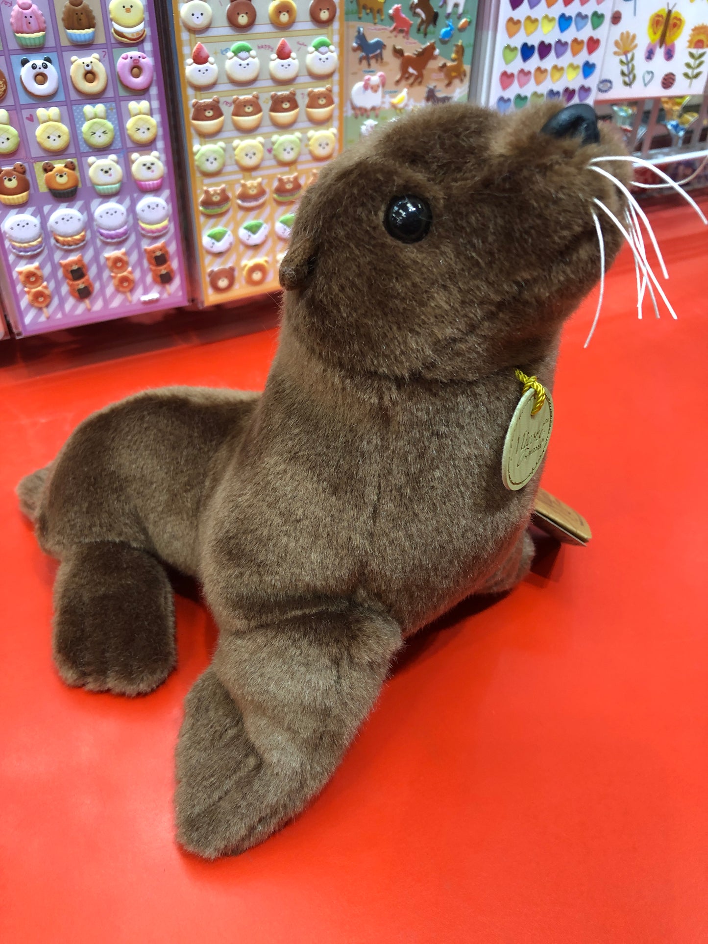 Realistic brown Aurora Miyoni sea lion plush toy with whiskers and tag, displayed on red surface with colorful stickers in background