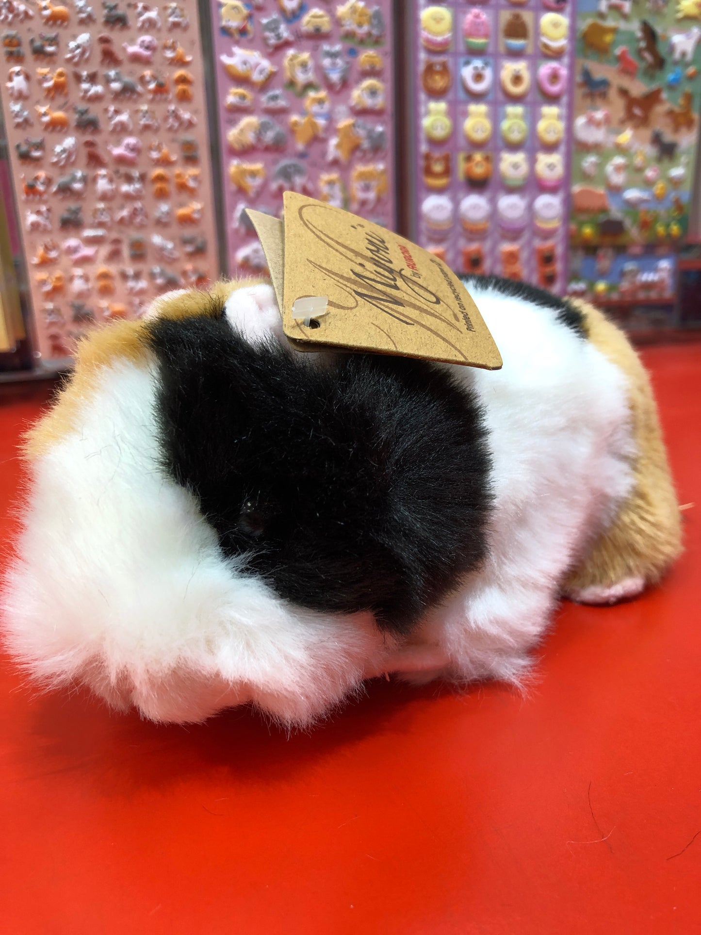 Realistic tri-colored American Guinea Pig plush toy with gold tag on red surface, colorful sticker sheets in background
