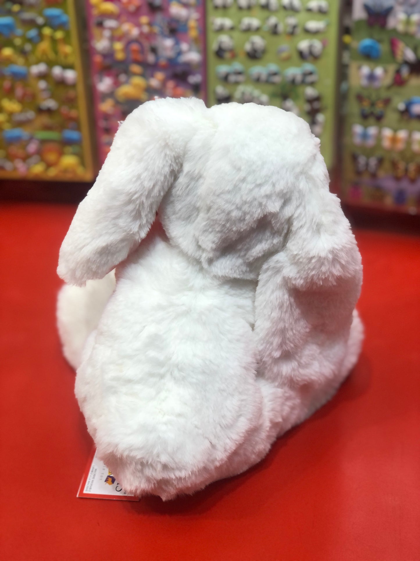 Soft white bunny plush toy with long ears, sitting on red surface, colorful toy display in background