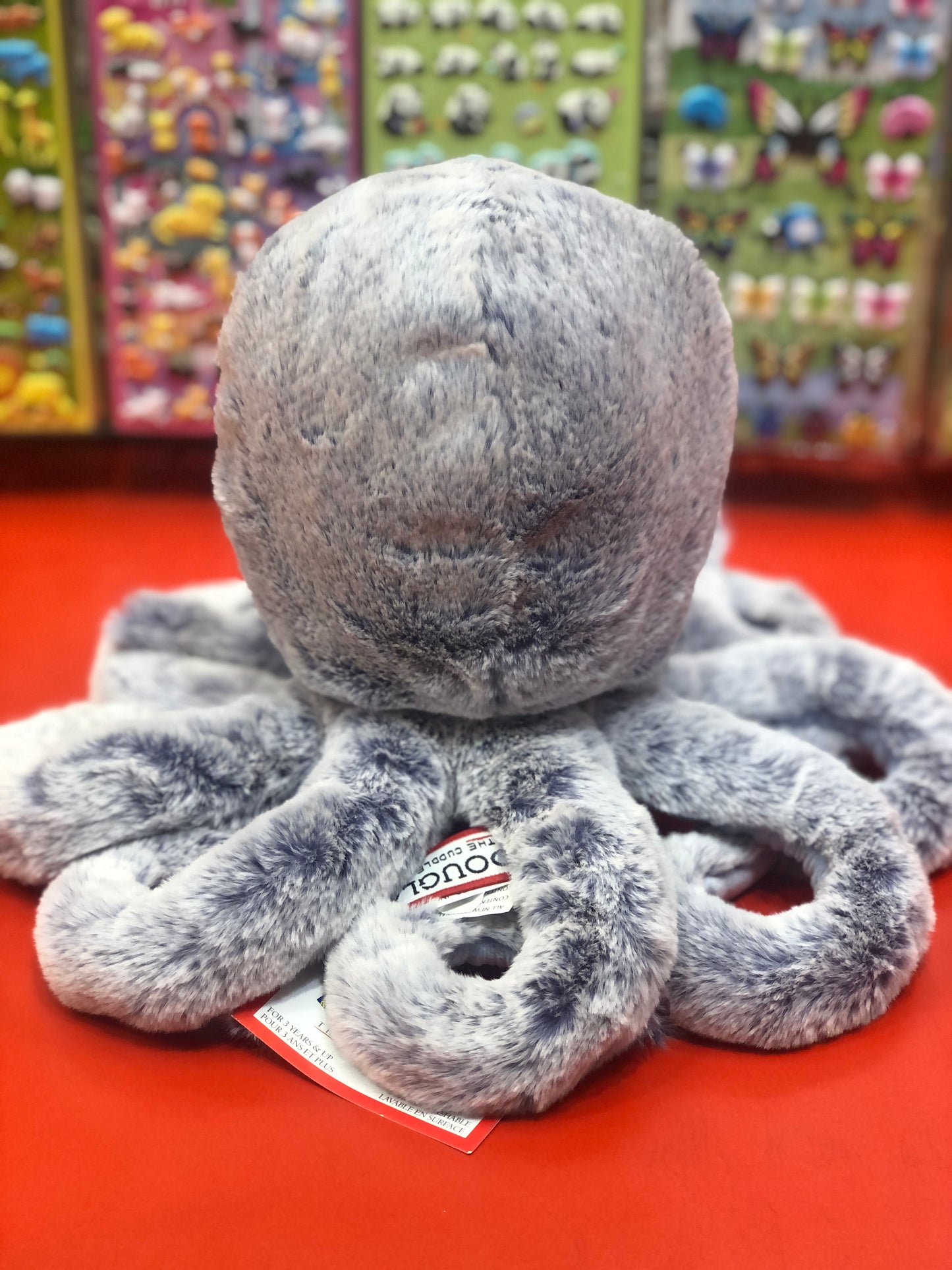 Soft lavender octopus plush toy with frosted tips and periwinkle tentacles on red display, colorful toy shelves in background