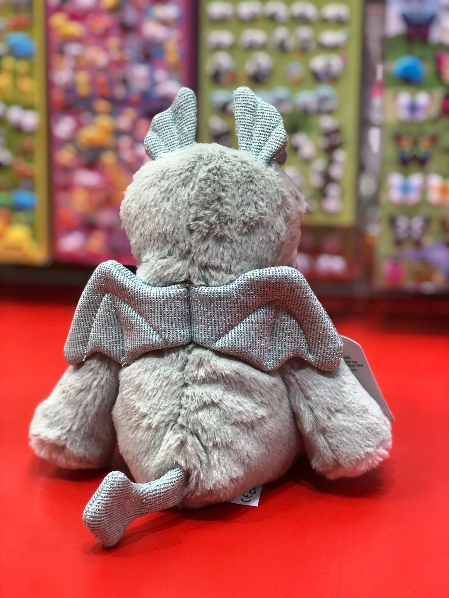 Soft gray Marshmallow Zoo dragon plush toy with sparkly wings and tail, displayed on red surface with colorful toy backdrop