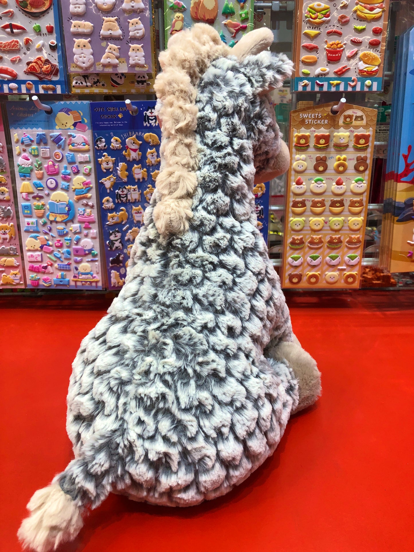 Soft plush giraffe toy with neutral-colored fur displayed in front of colorful sticker sheets in a toy store