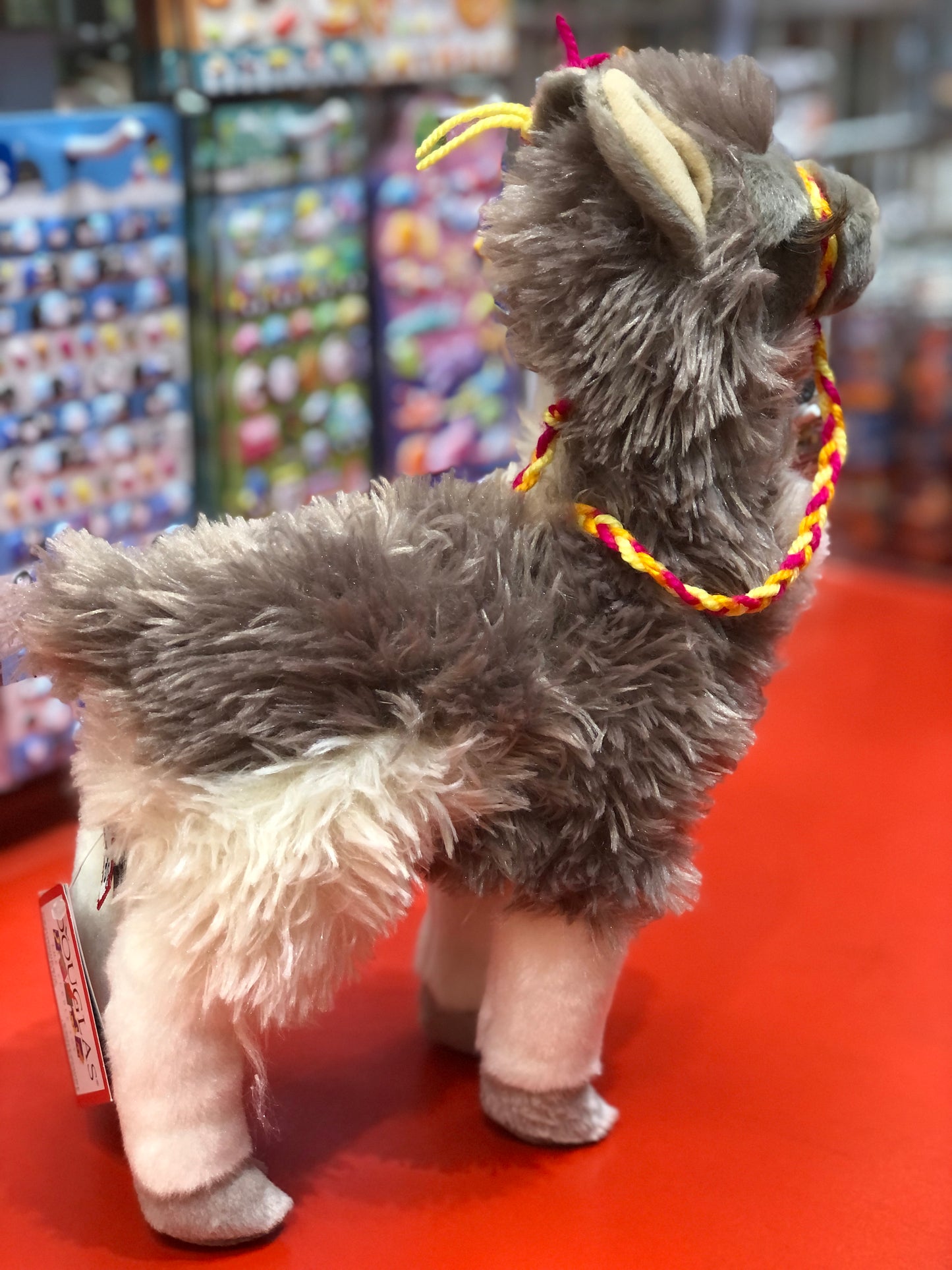 Plush Douglas Zephyr Llama toy standing upright, with gray fluffy fur and colorful harness, displayed in toy store