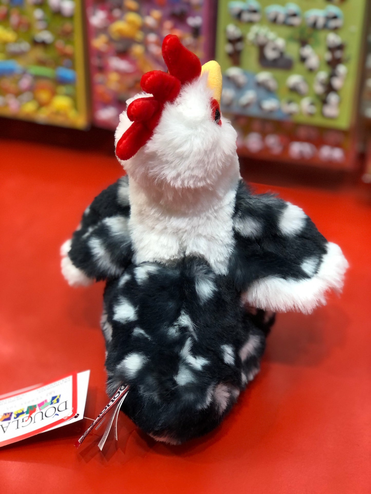 Colorful Douglas Romie Rooster plush toy with red comb, white head, and spotted black body on display in toy store