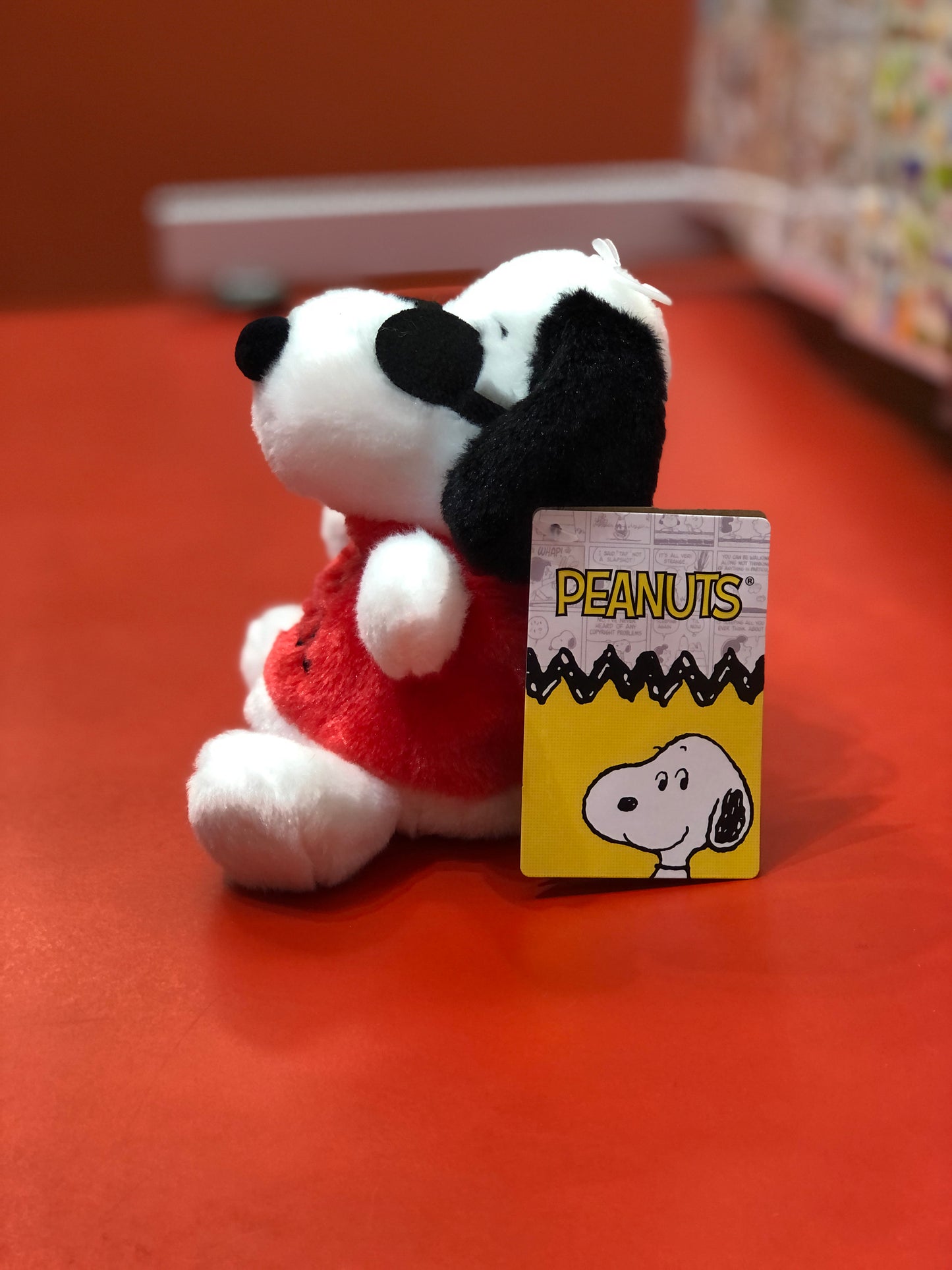 Aurora Peanuts Joe Cool plush toy with red outfit, seated next to official Peanuts tag featuring Snoopy on orange surface