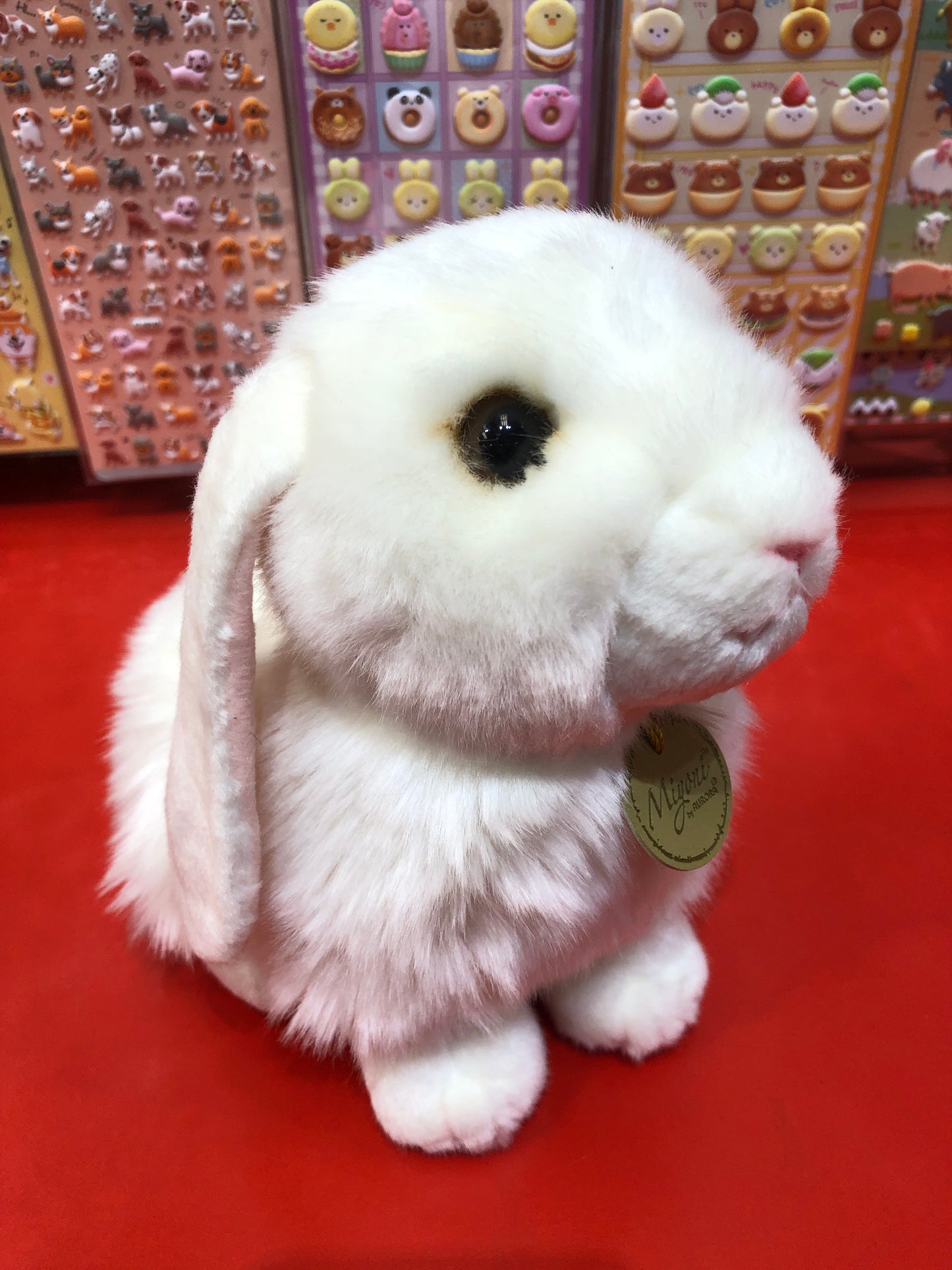 Adorable white lop eared bunny plush with fluffy fur and long ears, sitting on red surface with colorful stickers in background