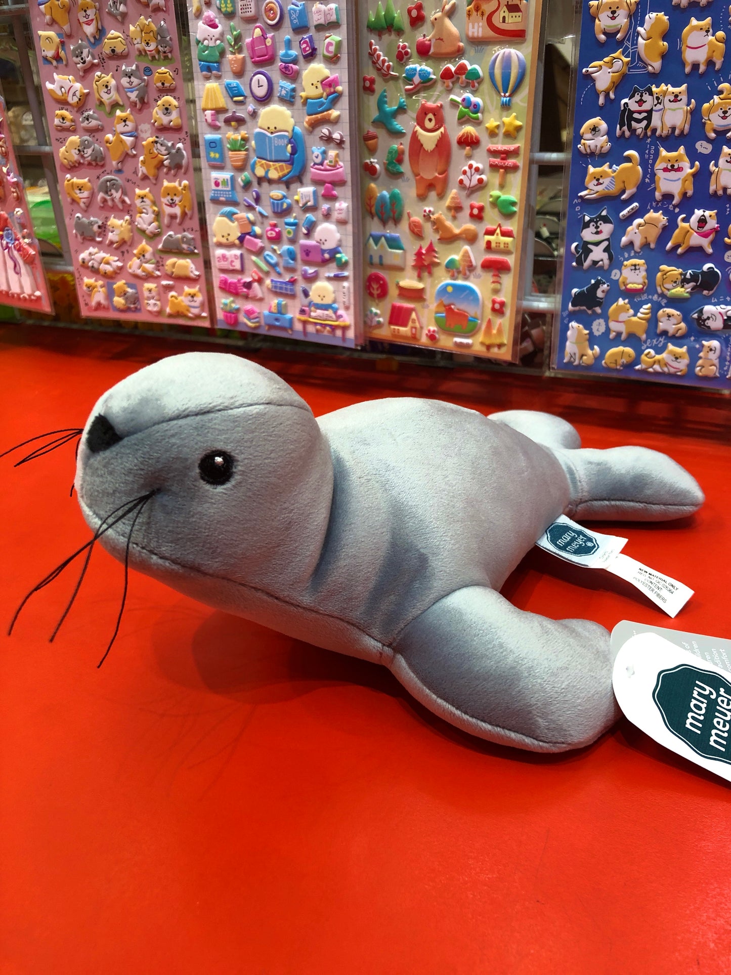 Soft gray Mary Meyer Smootheez Seal plush toy on red display, with colorful sticker sheets in background