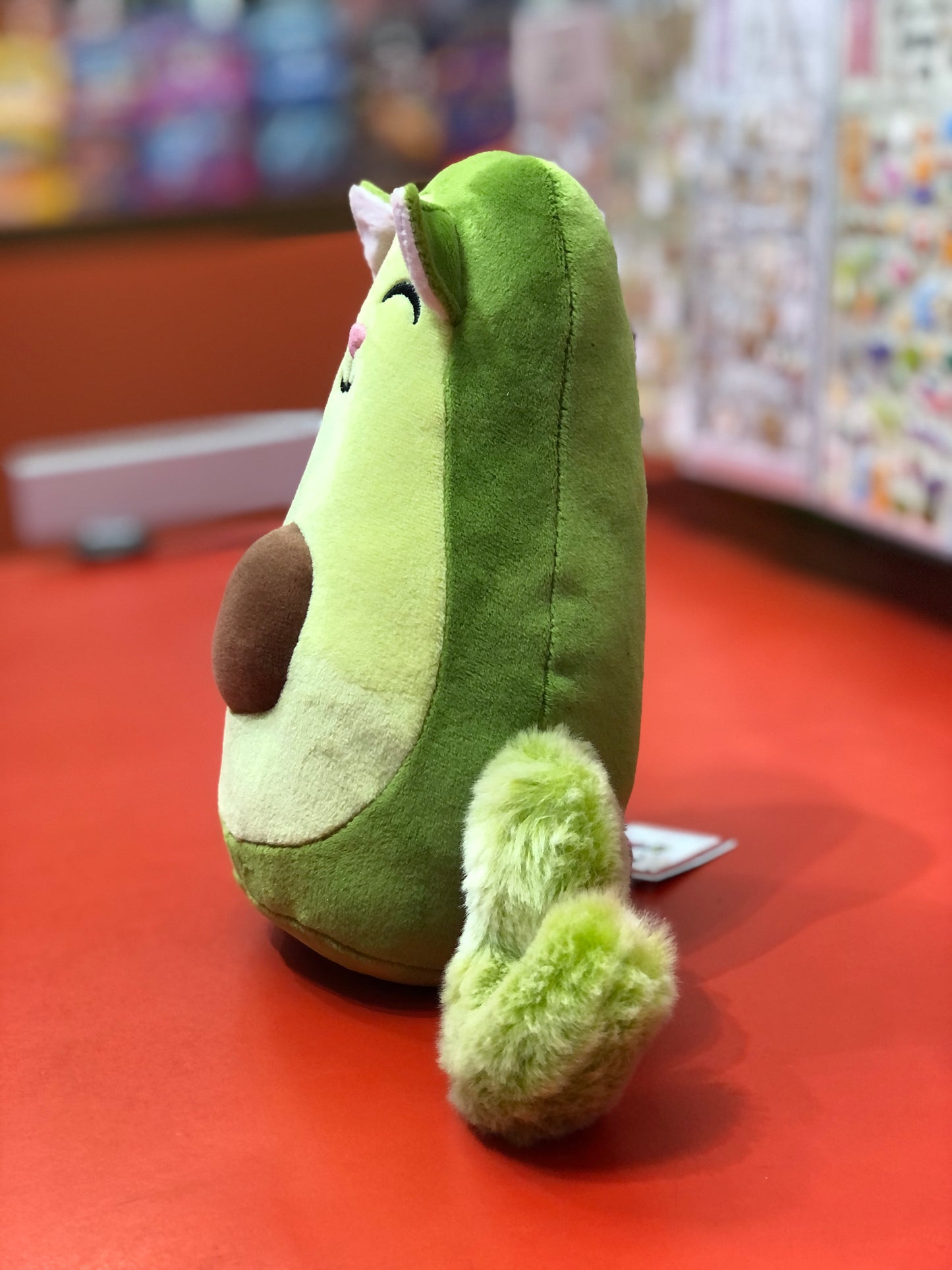 Whimsical Avogato plush toy combining avocado and cat, with green microfiber body, brown pit, and fluffy tail on red surface