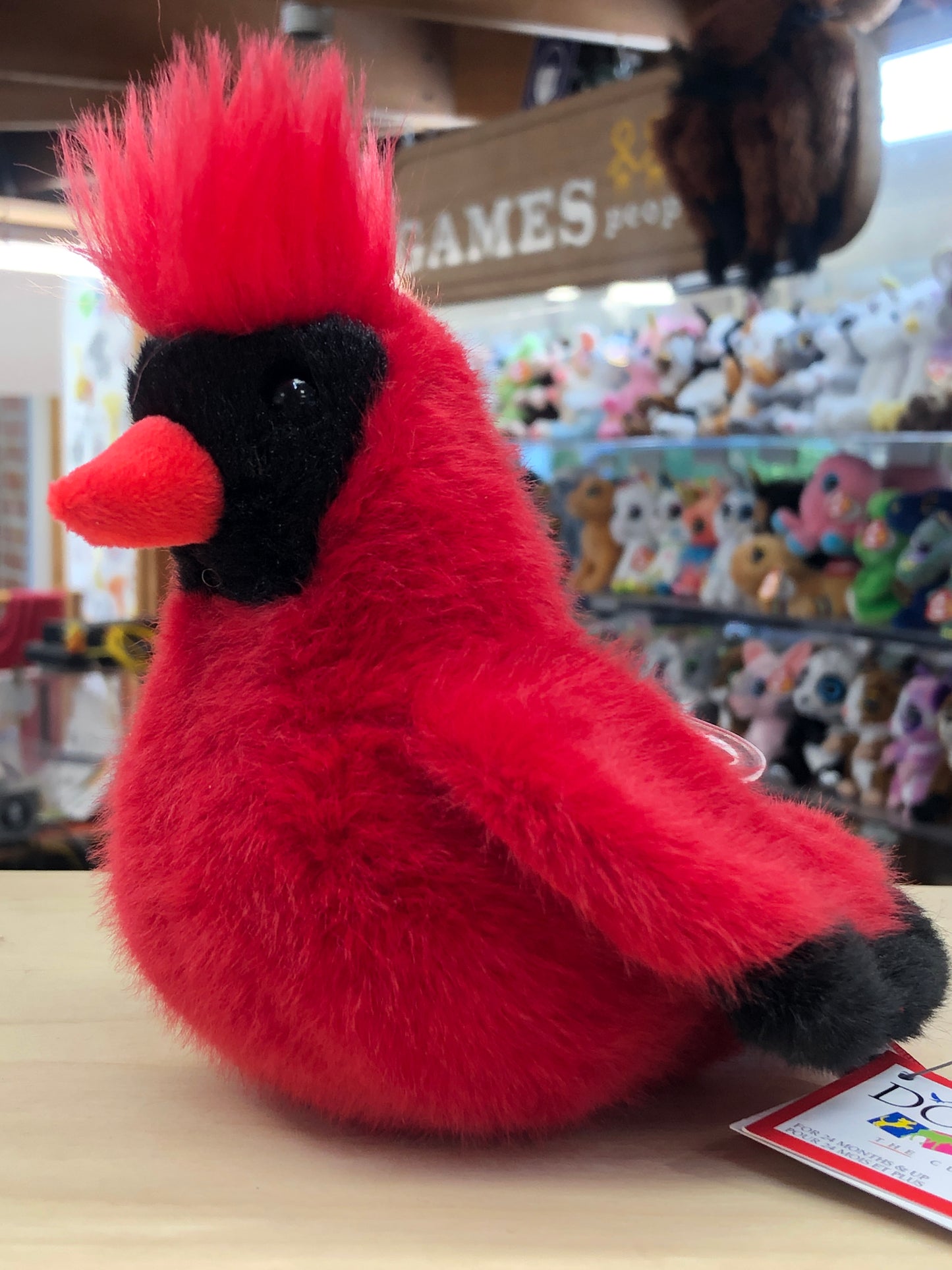 Soft, vibrant red cardinal plush toy with black facial markings and tufted crest, displayed in a toy store among other stuffed animals