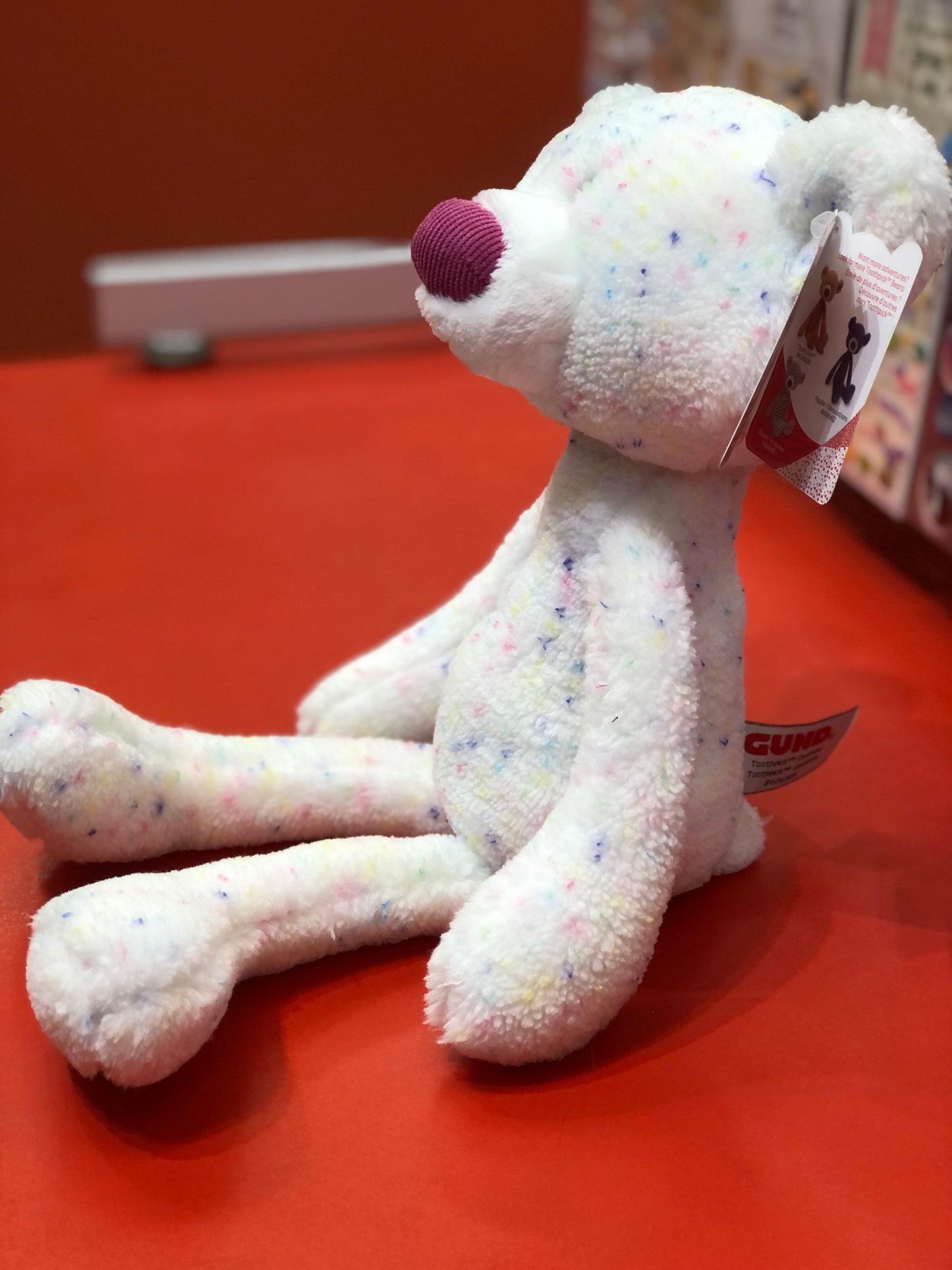 Gund Toothpick Confetti Bear plush toy with colorful speckles and purple nose, sitting on red surface