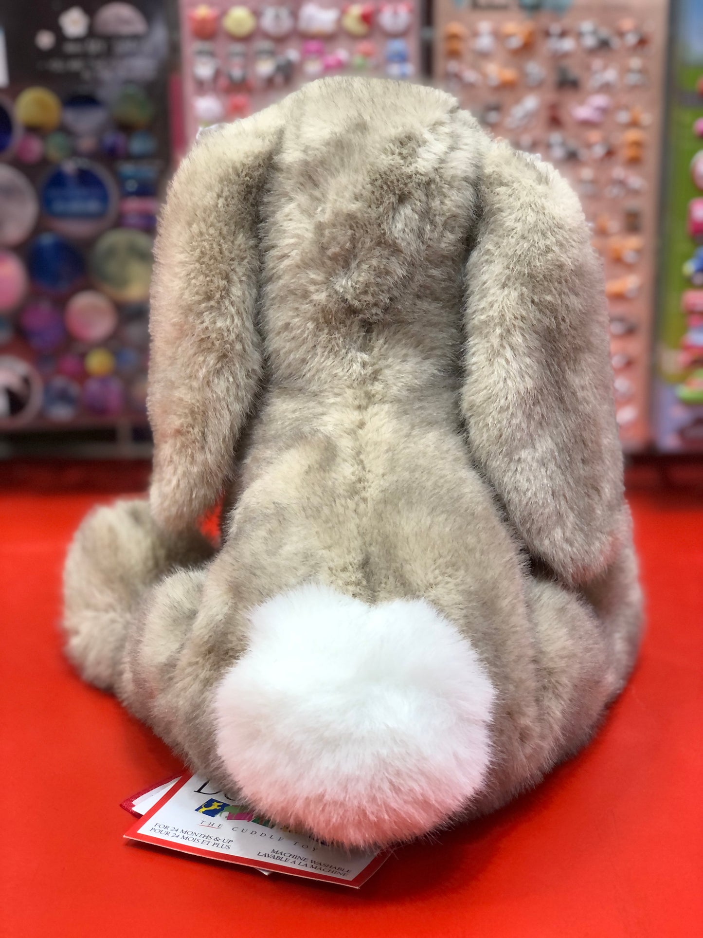 Soft plush Douglas Toastie Bunny toy, 8 inches tall, with fluffy gray fur and white tail, sitting on red surface in store display