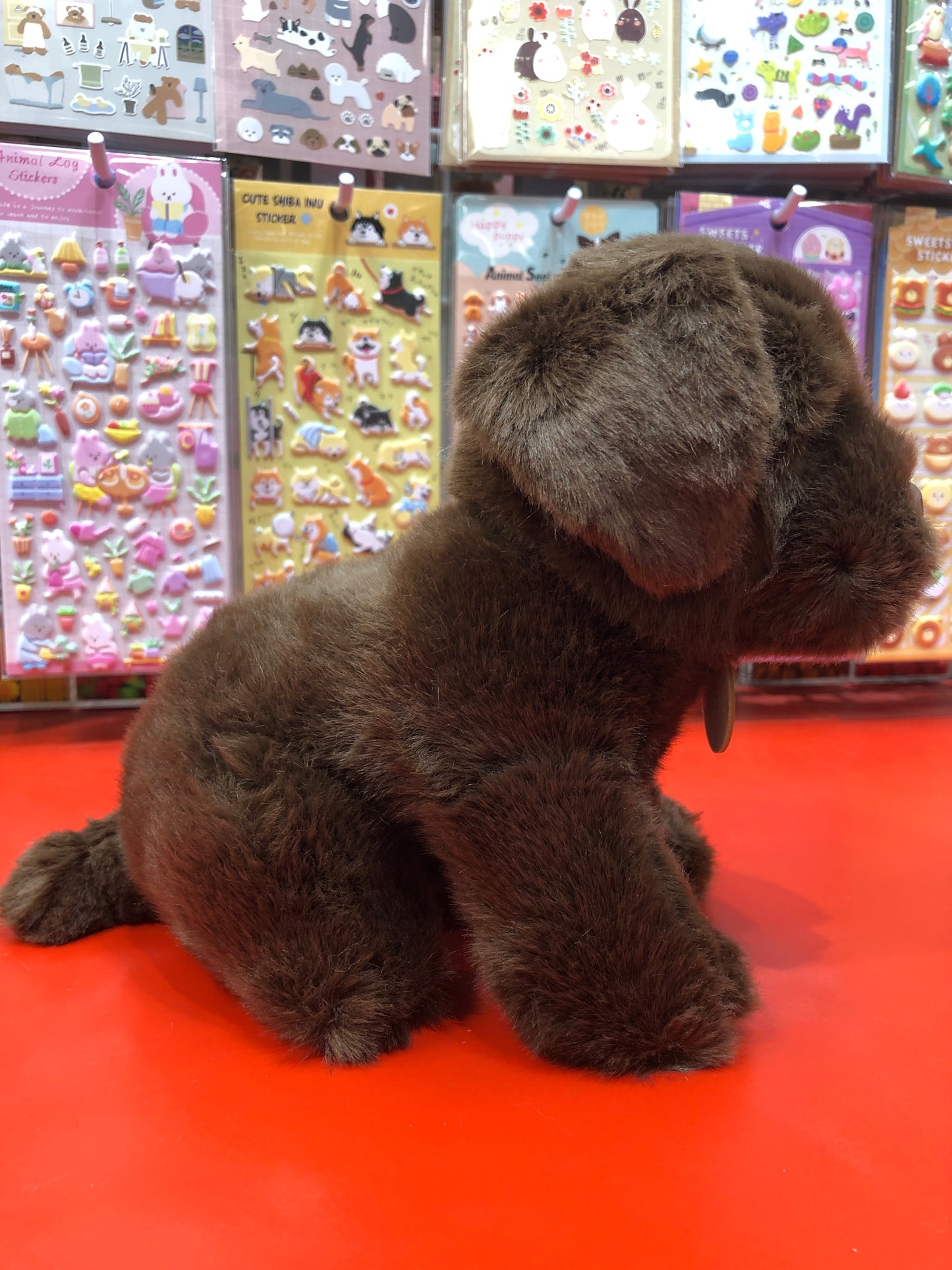 Plush chocolate Labrador puppy sitting on red surface with colorful sticker displays in background