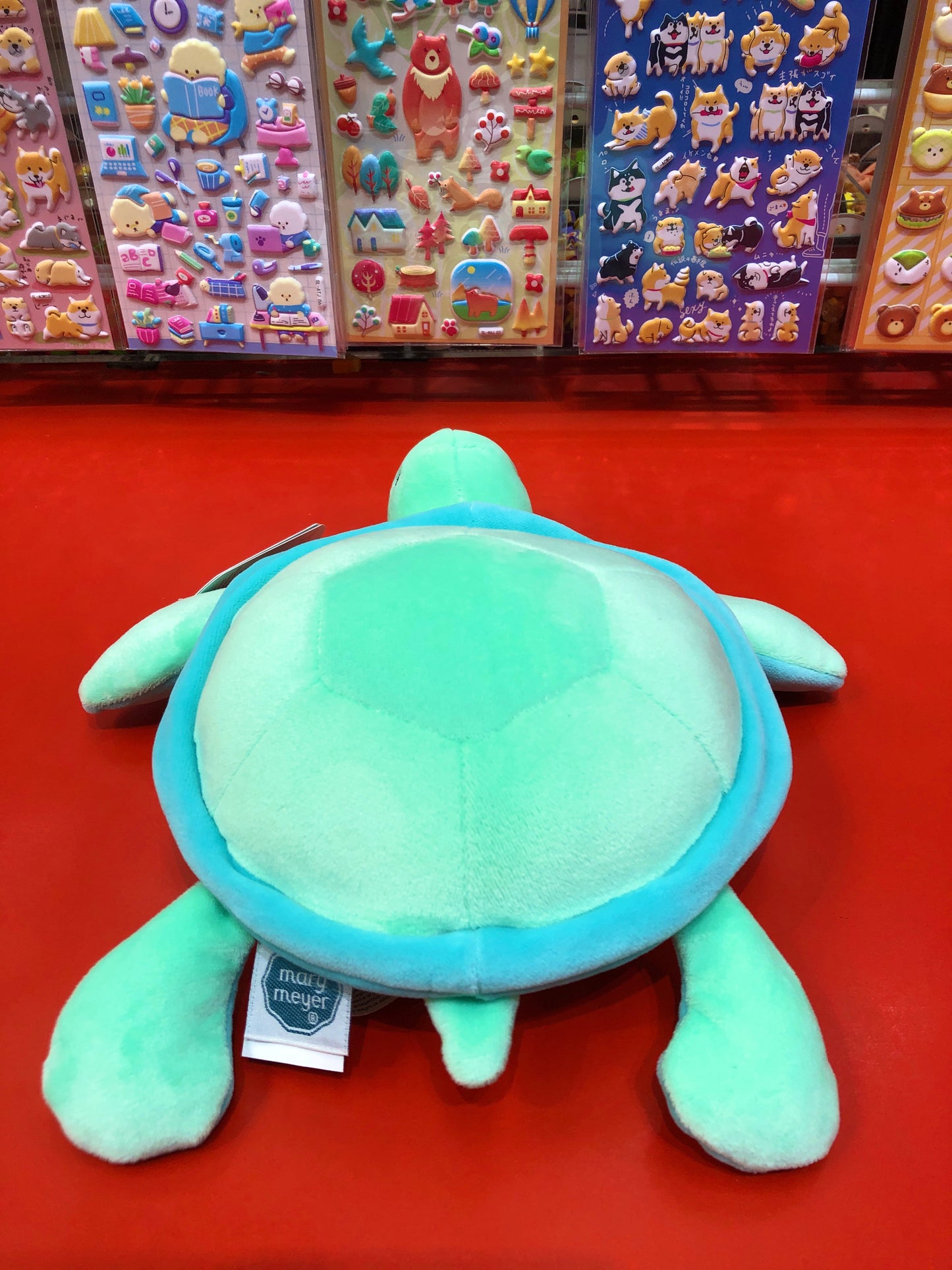 Mary Meyer Smootheez sea turtle plush toy on red surface, colorful sticker sheets in background, soft and squeezable design
