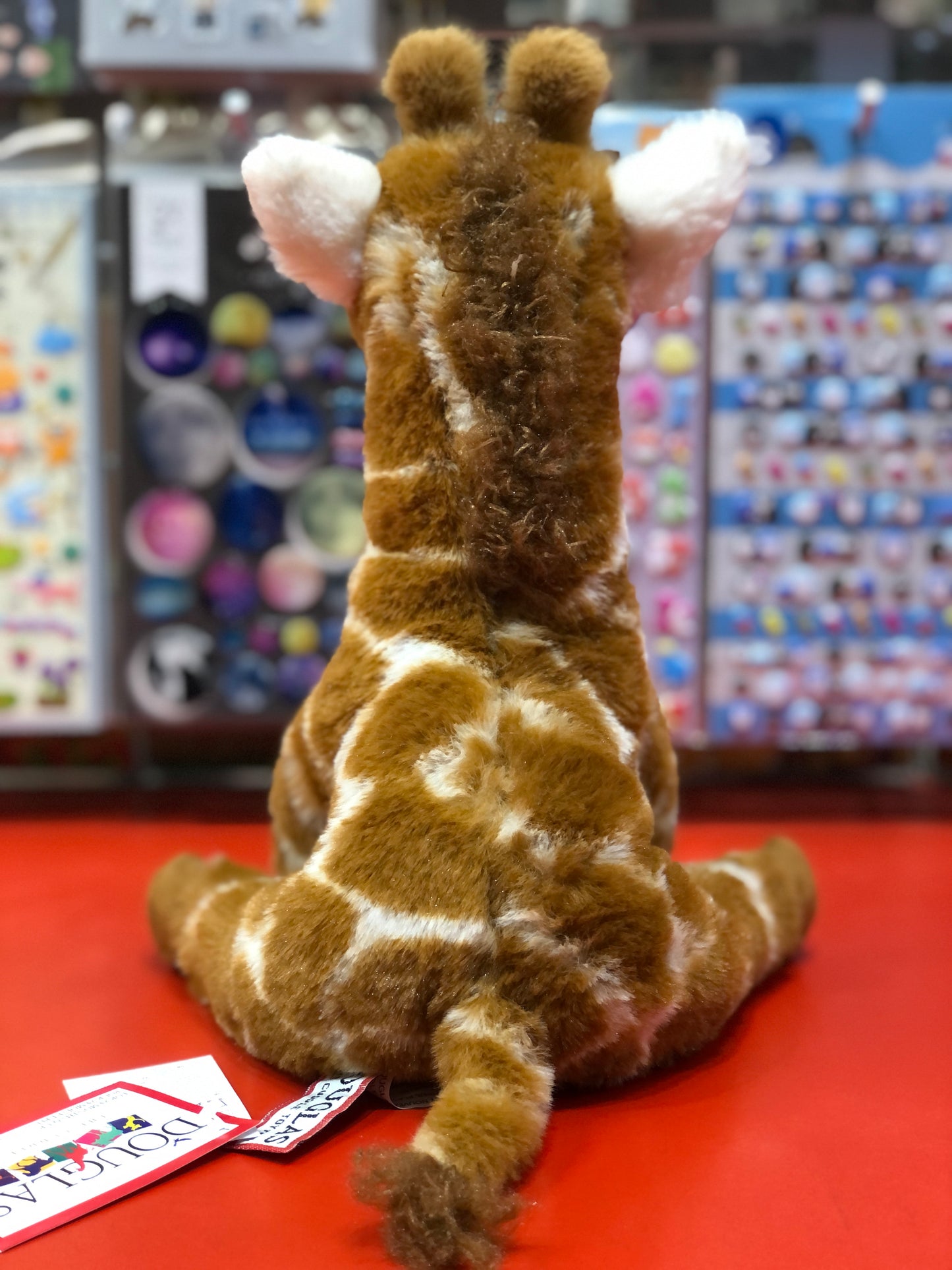 Soft plush giraffe toy Jessie with spotted coat, sitting on red surface in toy store display, back view showing detailed design
