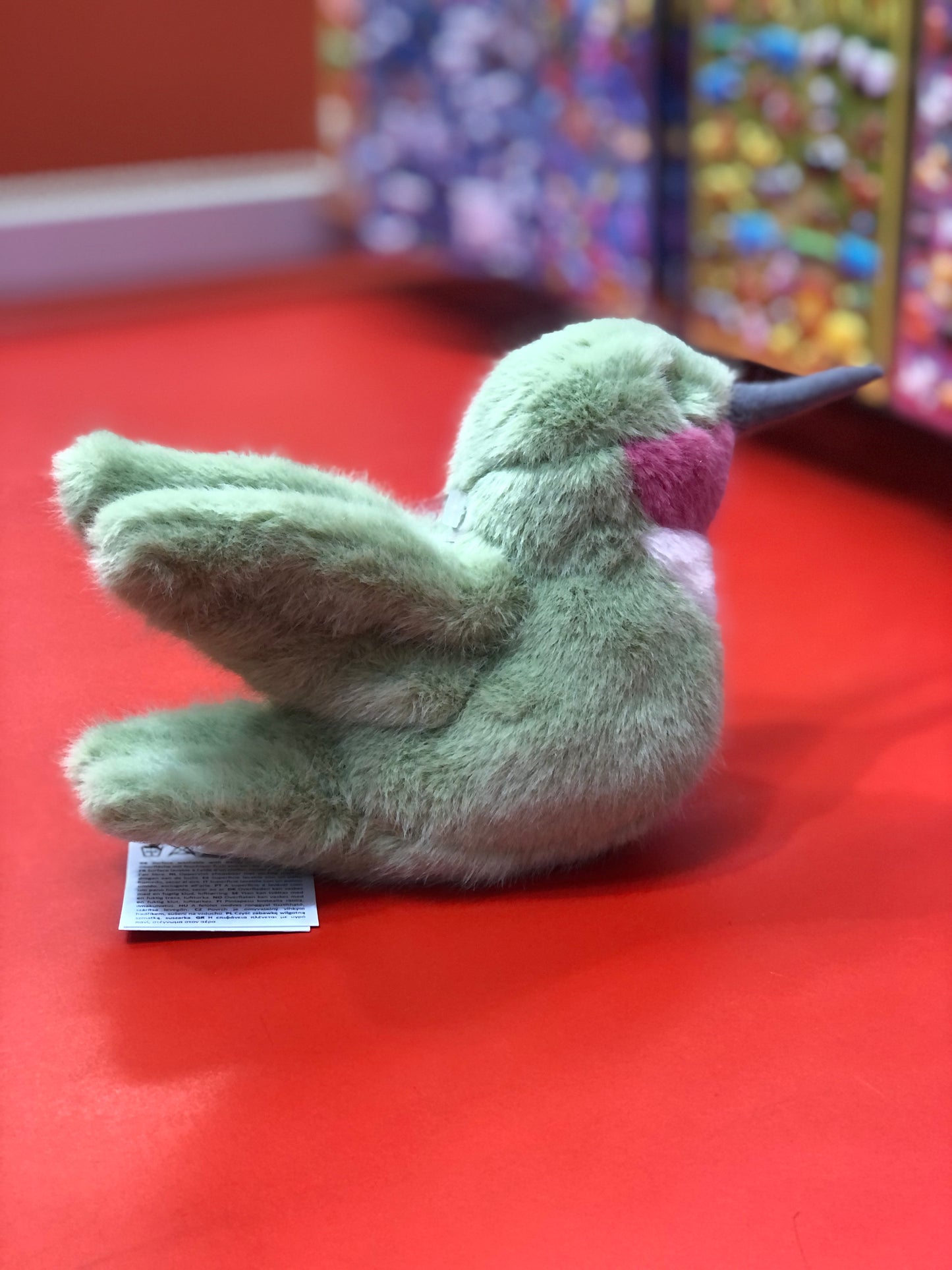 Soft green plush hummingbird toy with rosy throat on red surface, showcasing delicate features and lifelike design