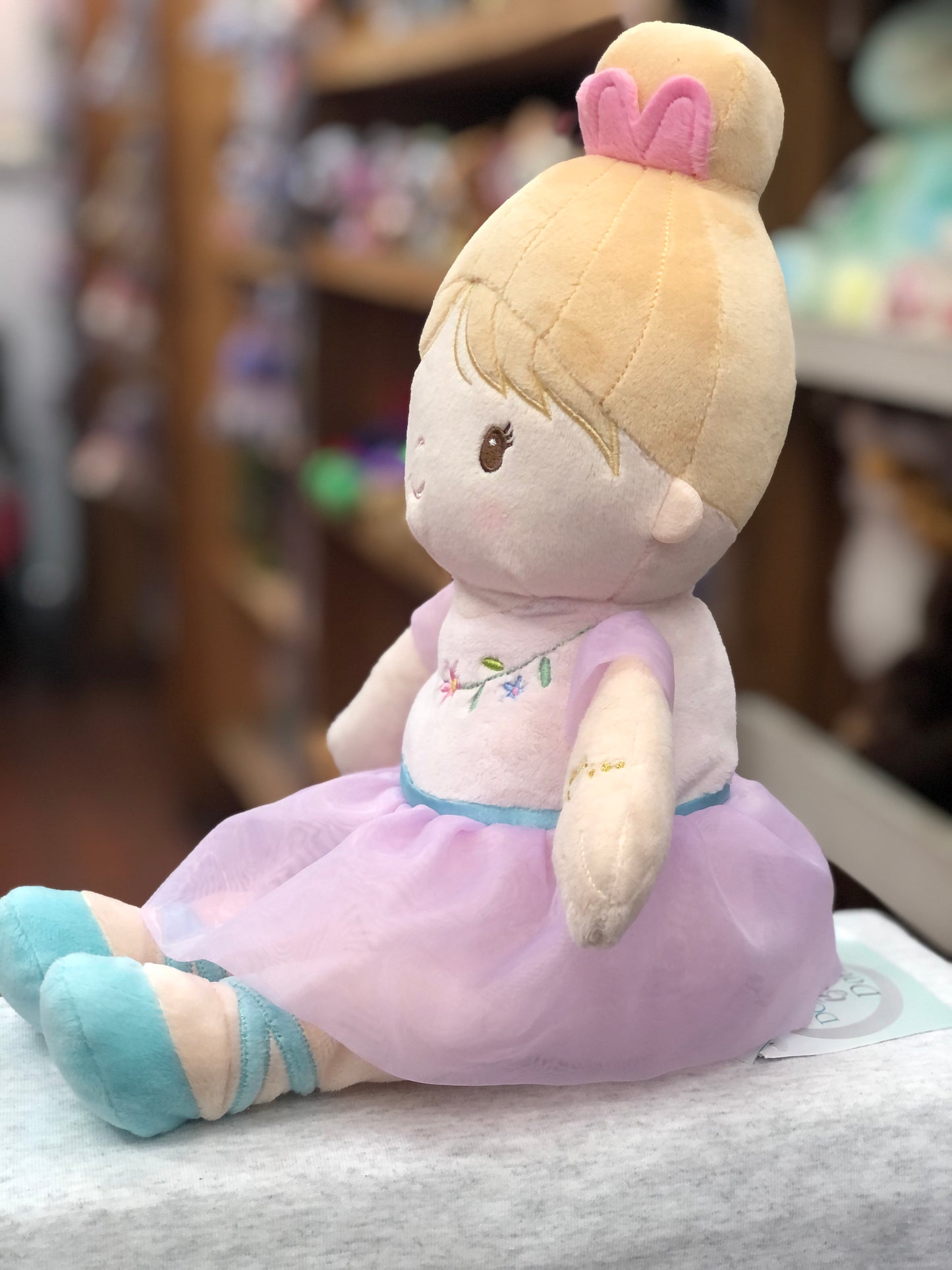 Soft plush ballerina doll with blonde hair bun, pink tutu, and blue ballet slippers sitting on display in toy store
