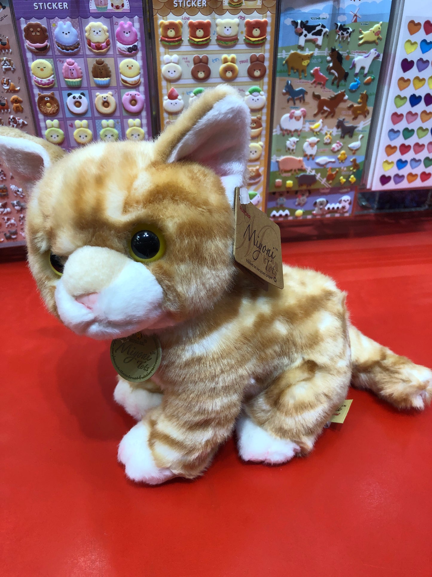 Adorable Aurora Miyoni orange tabby kitten plush toy with realistic features, displayed on red surface near colorful sticker sheets