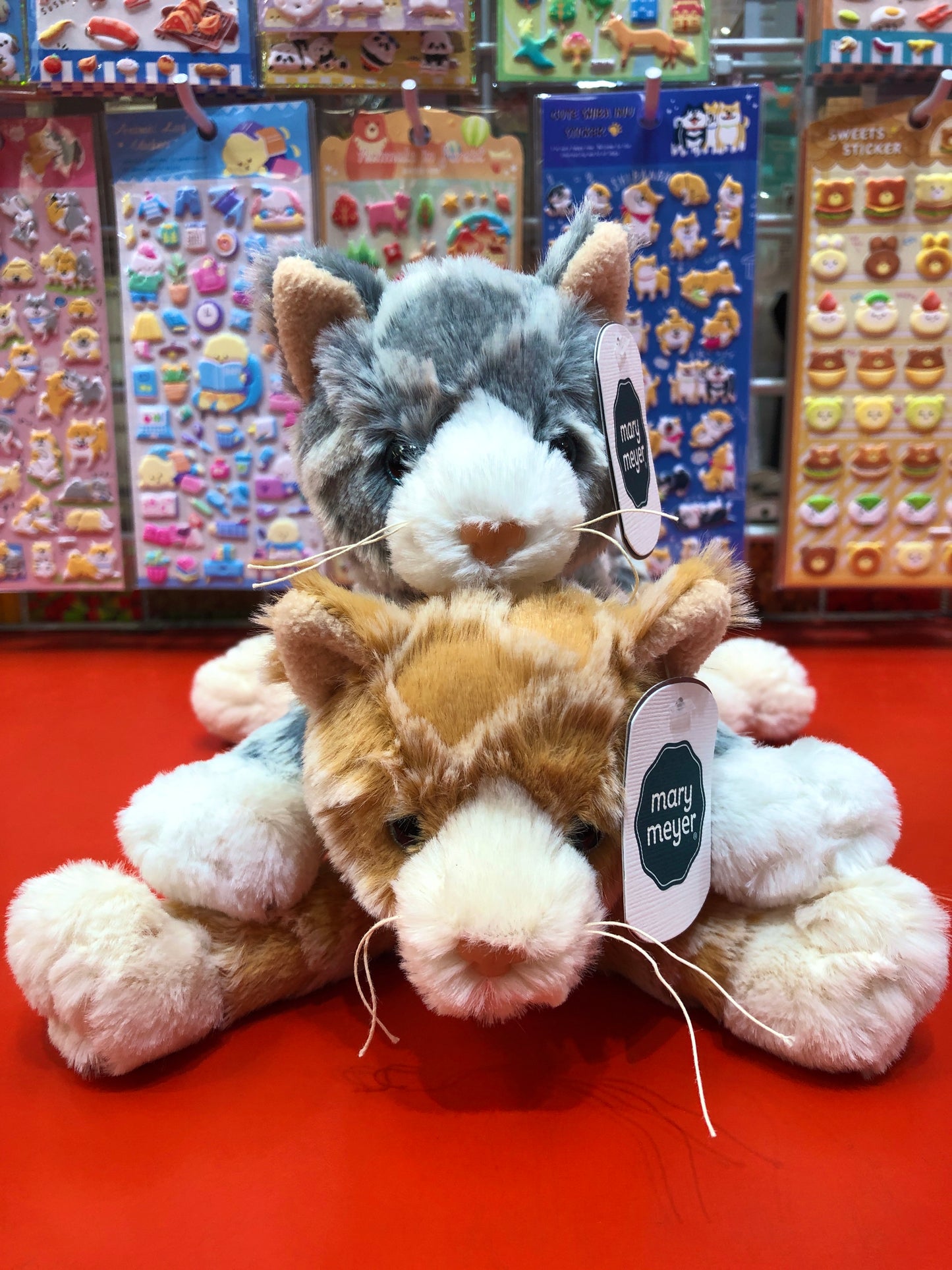 Mary Meyer FabFuzz Waffles Kitten plush toys in gray and orange, displayed with colorful sticker backgrounds in toy store