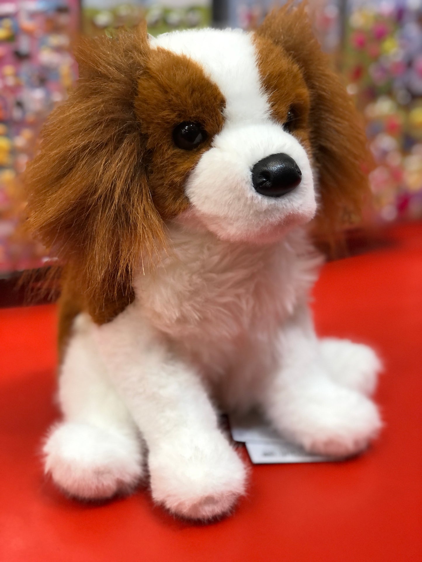 Adorable Cavalier King Charles Spaniel plush toy with chestnut and white fur, sitting on red surface, large eyes and floppy ears