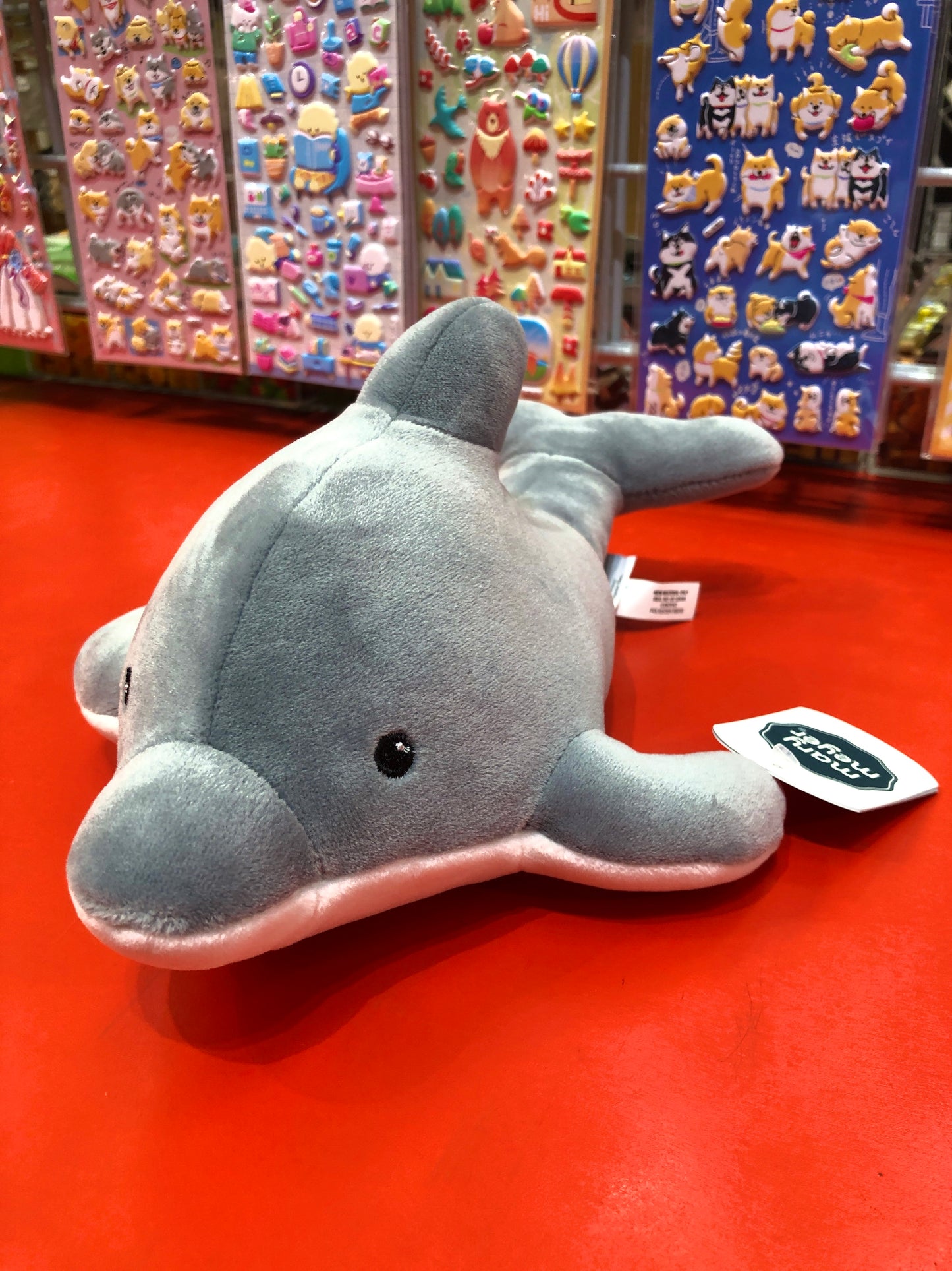 Soft gray Mary Meyer Smootheez dolphin plush toy on red display, with colorful sticker sheets in background