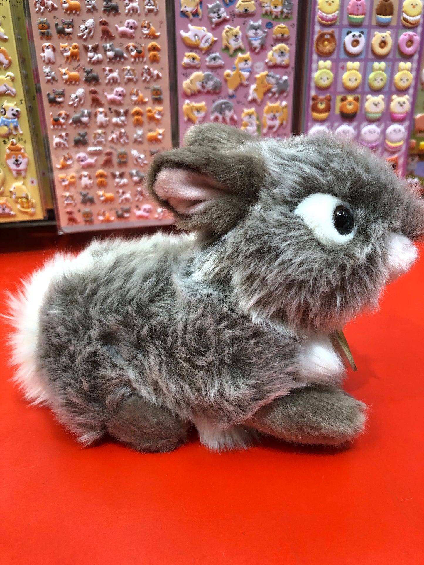 Adorable grey baby bunny plush toy with soft fur and bright eyes, displayed on red surface with colorful sticker sheets behind