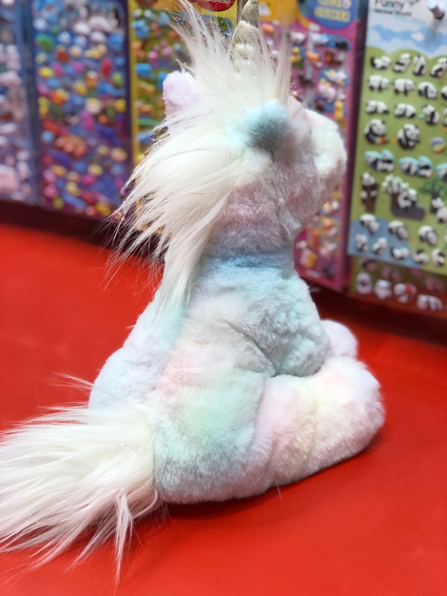 Soft rainbow unicorn plush toy with fluffy mane and tail, displayed on red surface with colorful toy packaging in background