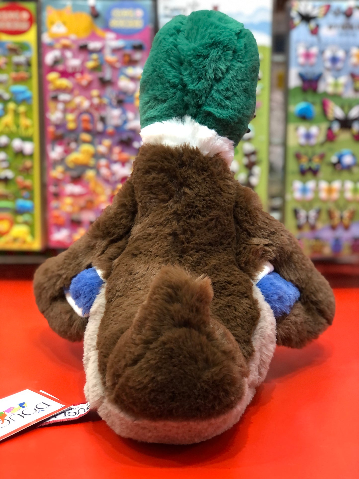 Soft mallard duck plush toy with green head, brown body, and blue wing patches on display in a colorful toy store