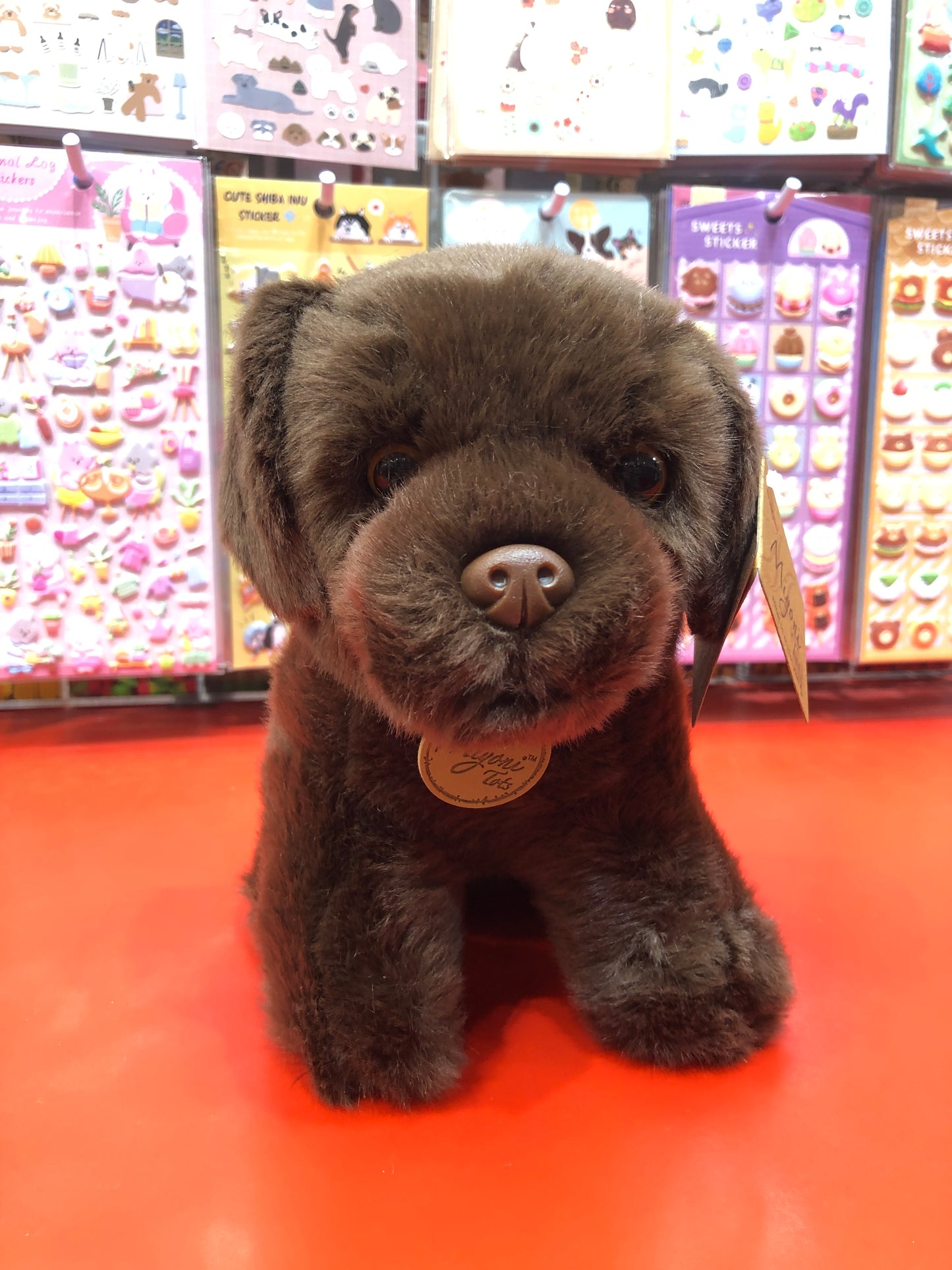 Adorable chocolate Labrador puppy plush toy with realistic features, sitting on red surface with colorful sticker display background
