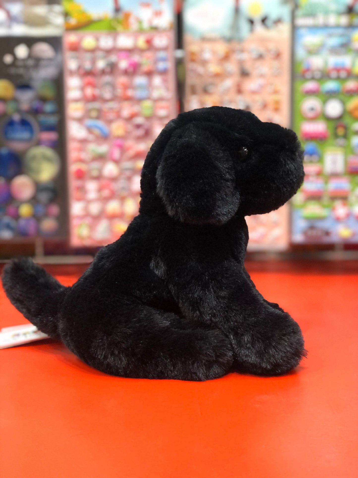 Cuddly mini black lab plush toy Hattie sitting on red surface, colorful toy display in background