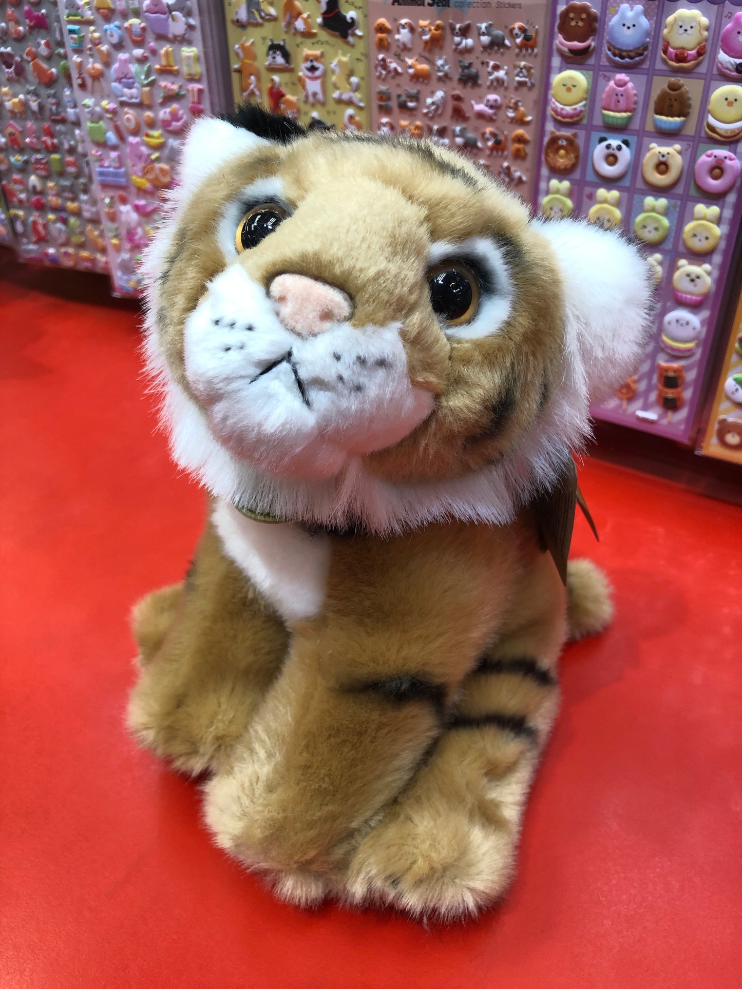 Adorable Bengal tiger cub plush toy with realistic features, sitting on red surface against colorful sticker background