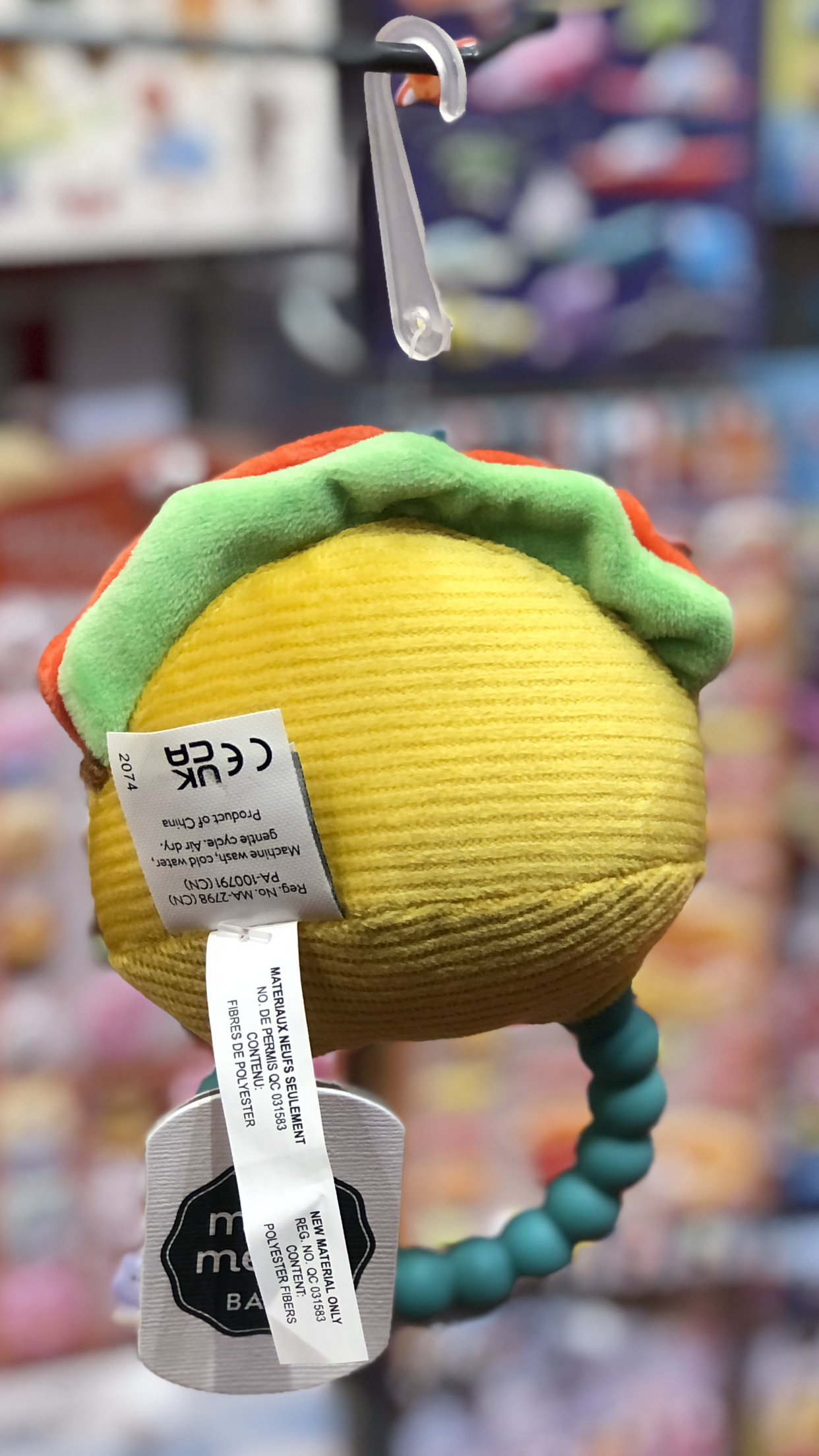 Mary Meyer taco-shaped teether rattle with soft yellow body, green and orange accents, hanging on display with product tags visible