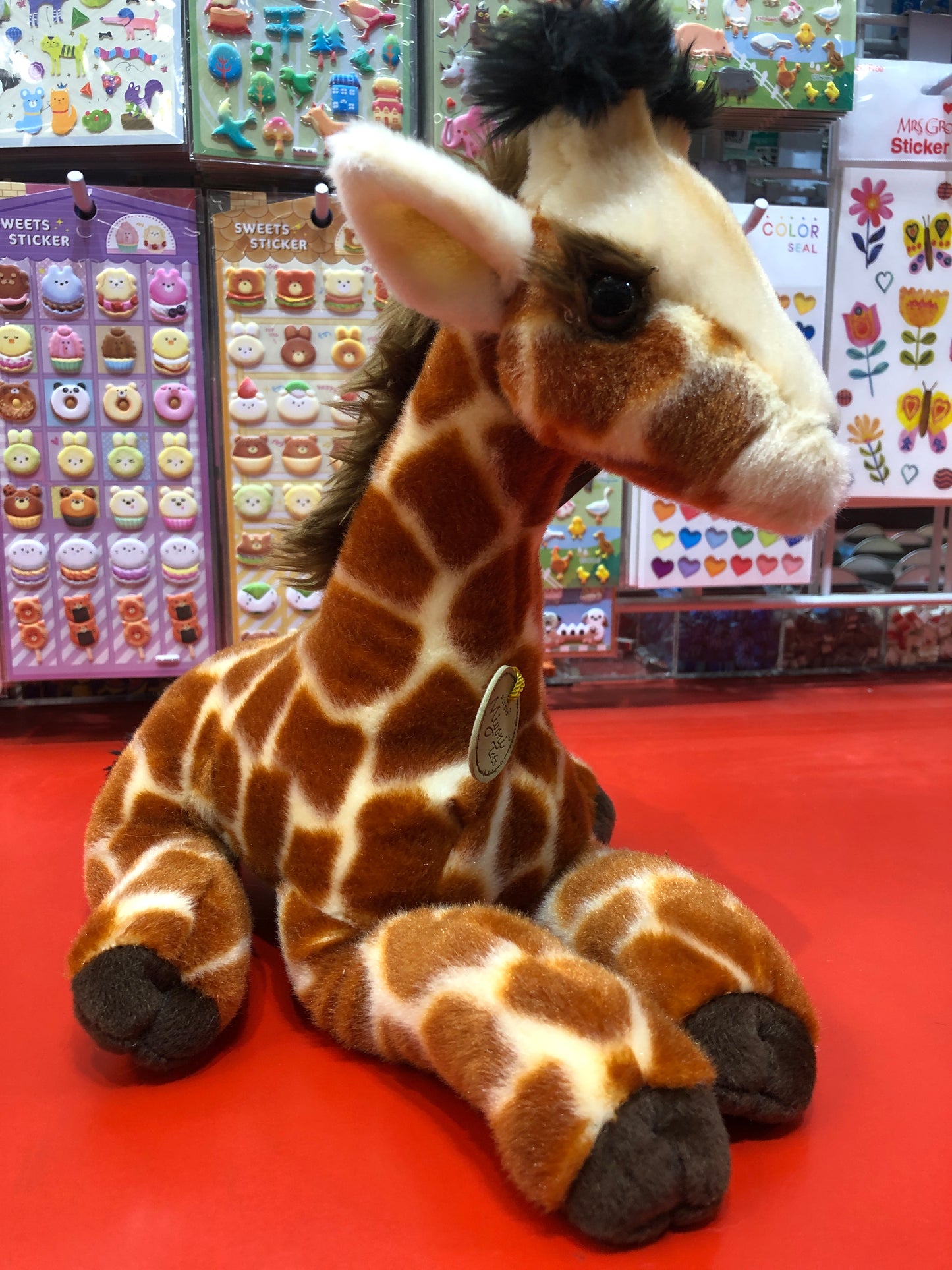 Adorable Aurora Miyoni Tots Giraffe Calf plush toy sitting on red display, surrounded by colorful sticker sheets in toy store