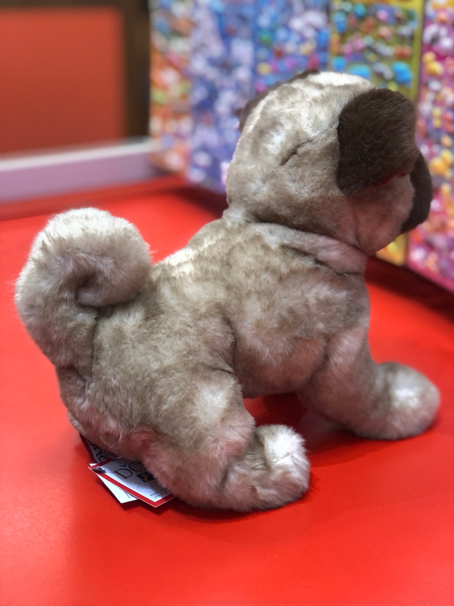 Soft, cuddly Milo Pug plush toy with curled tail and wrinkled features on red display, capturing breed's charm
