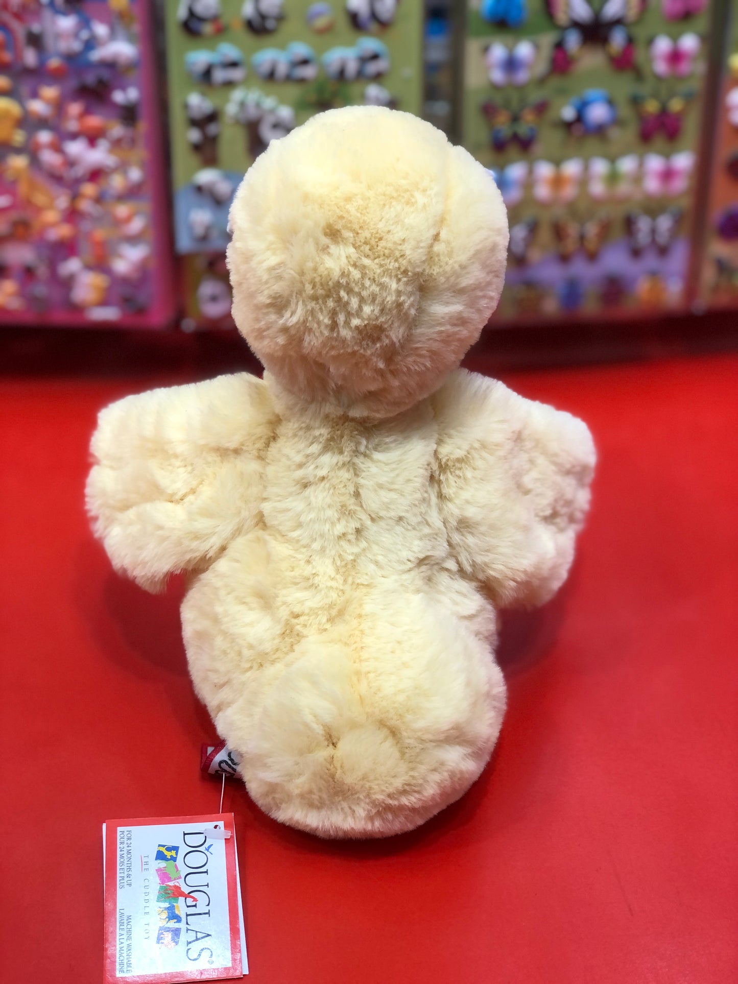 Soft yellow Douglas Mini Dennie duck plush toy sitting on red surface, with colorful plush display in background