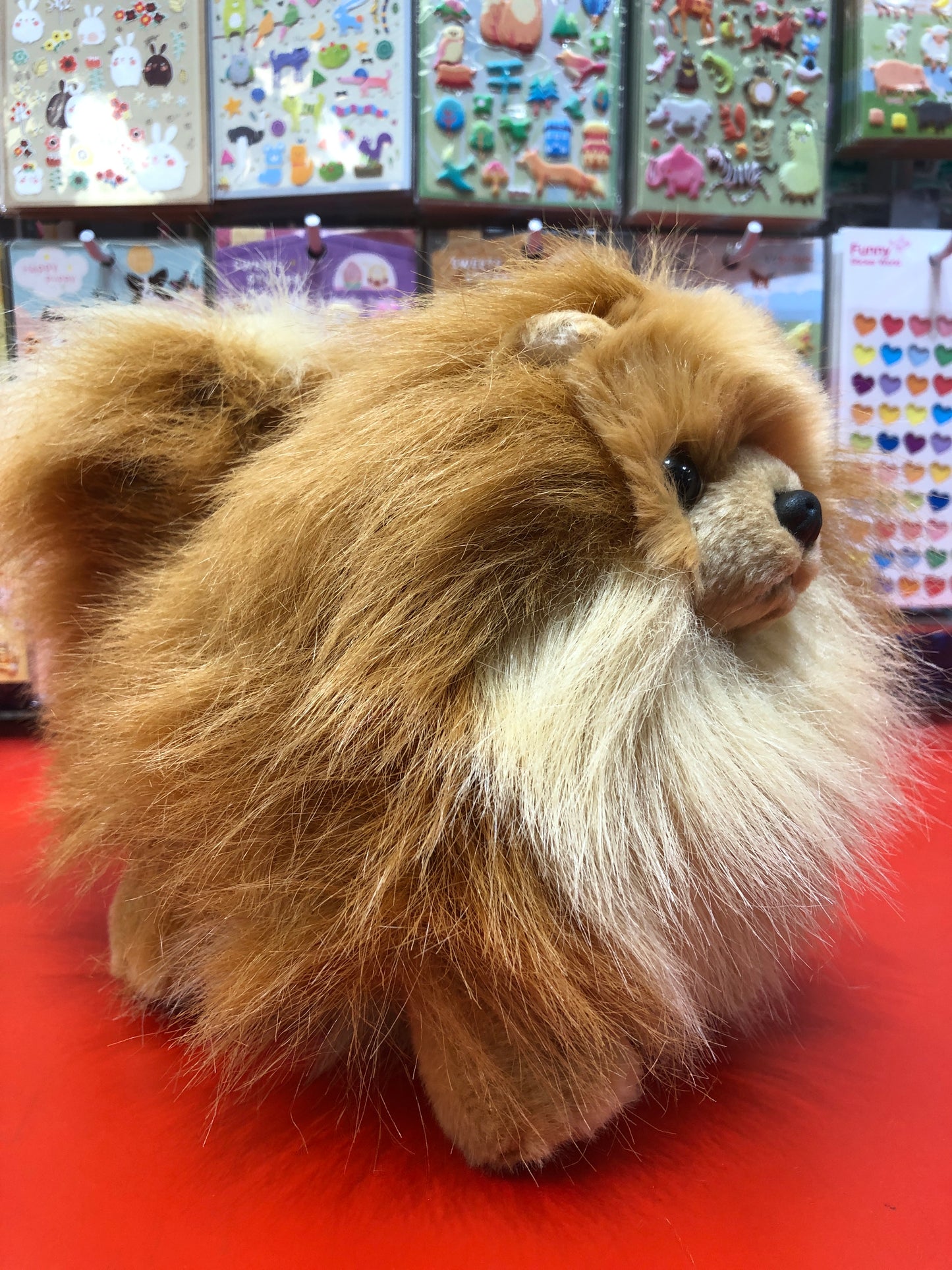Fluffy Pomeranian plush toy with tan and light brown fur on red display, colorful animal stickers in background