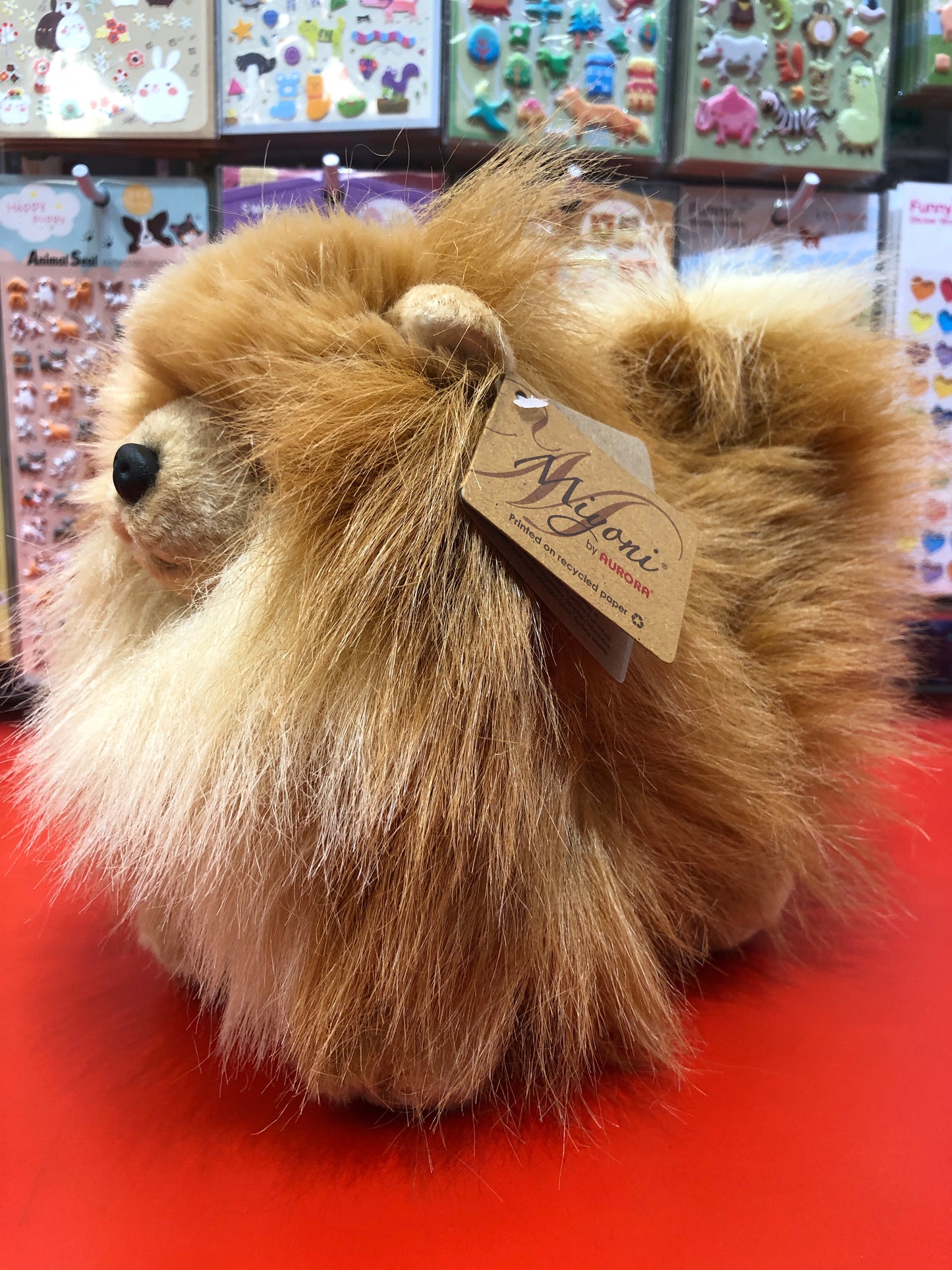 Fluffy Pomeranian plush toy with tan fur, displayed on red surface, tagged Miyoni by Aurora, surrounded by colorful stickers