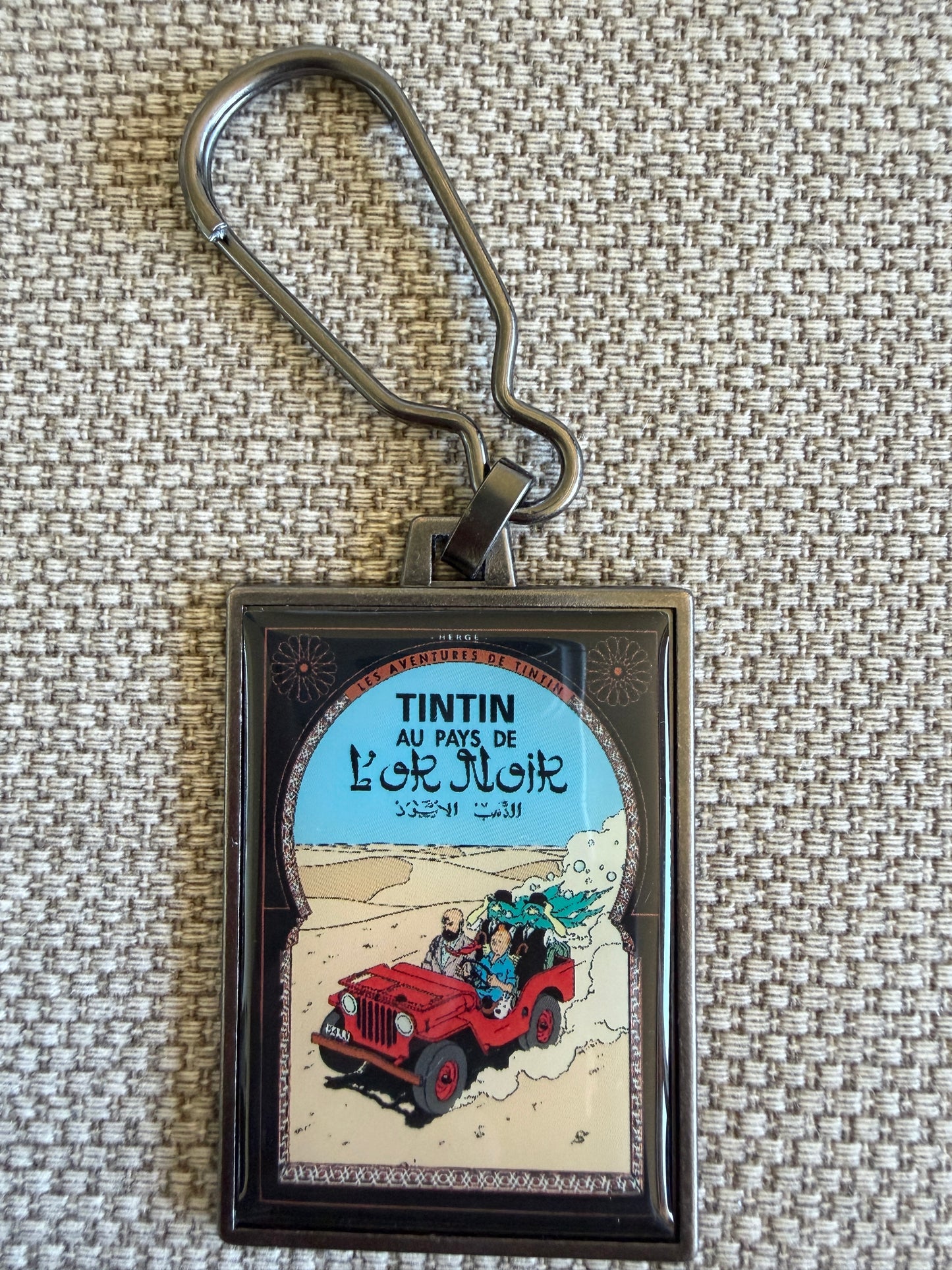A rectangular keychain displaying the French book cover art for Hergé's Tintin adventure "Au Pays de l'or Noir." The illustration shows Tintin, Snowy, and Prince Abdullah speeding across sun-drenched desert dunes in a classic red Willys Jeep under a bright blue sky.